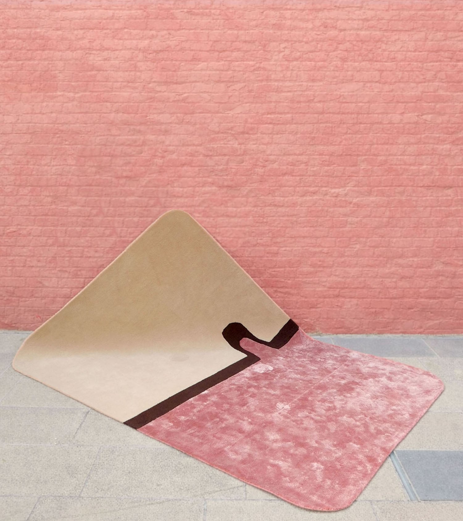 A geometric rug with beige and pink sections, featuring a black line, is partially folded against a pink brick wall on a light-colored floor.
