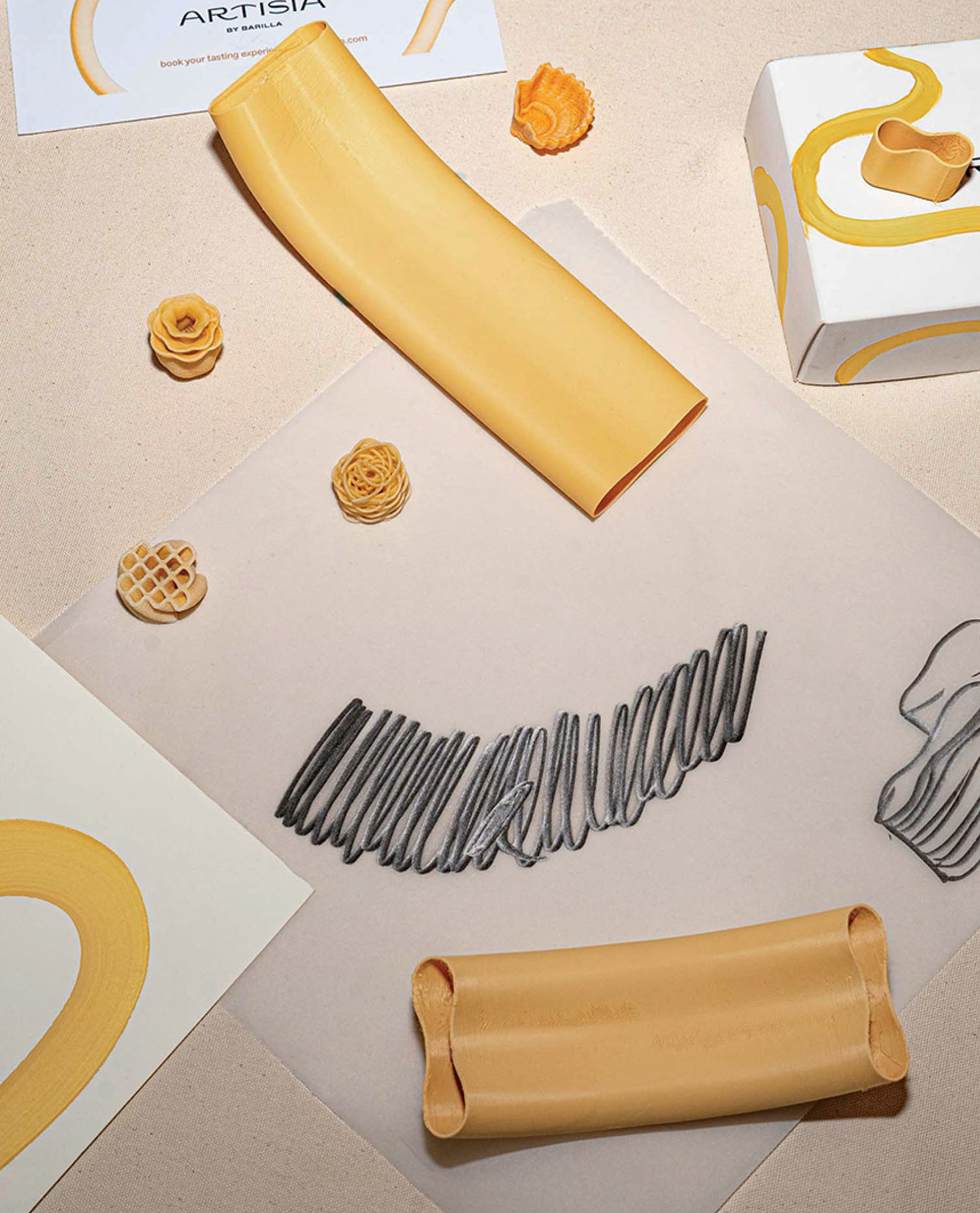 A top view of various uncooked pasta shapes, including flat, ridged, and rolled varieties, arranged on a light surface with papers and boxes featuring yellow and black brushstroke designs.