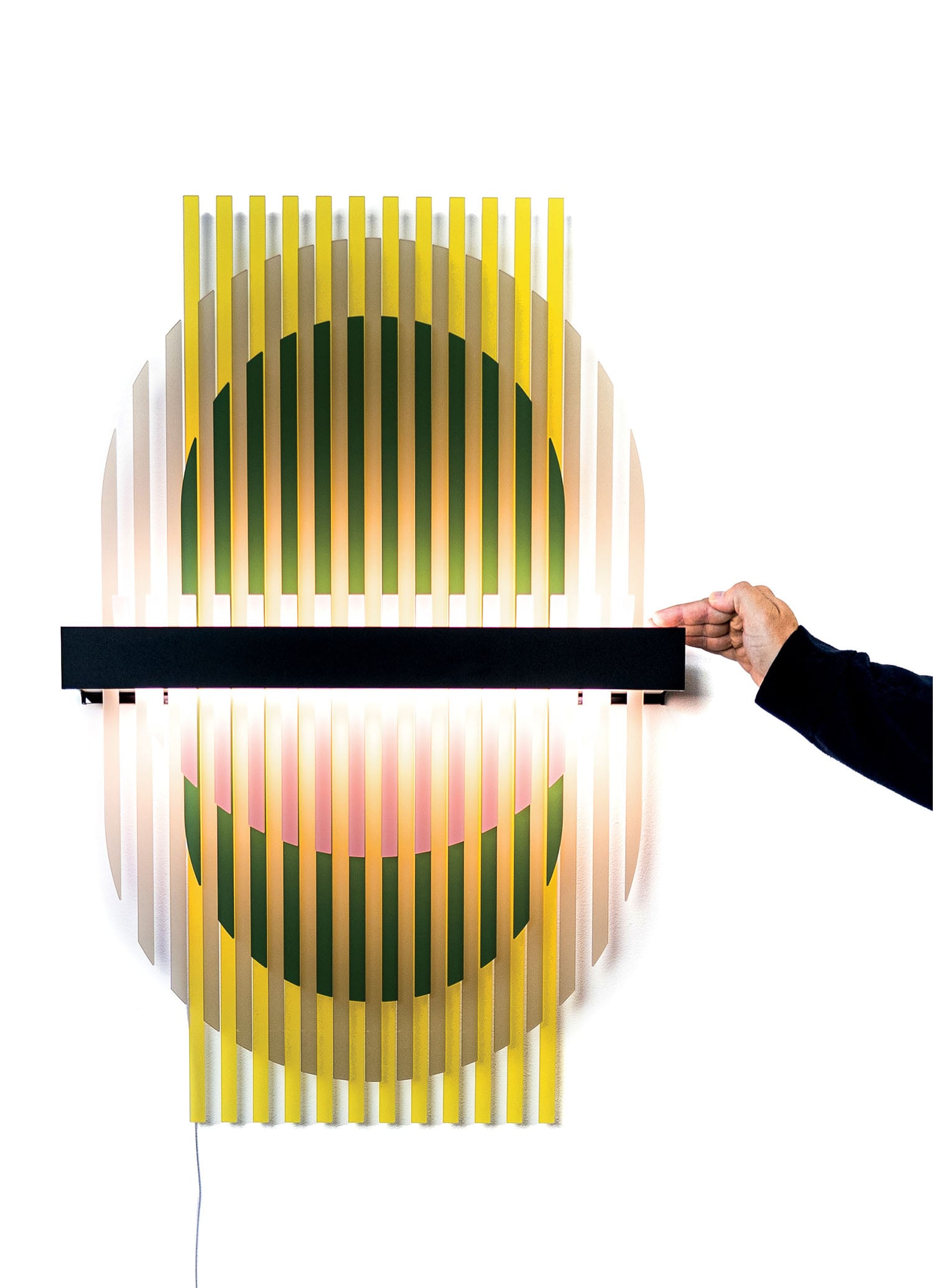 A hand adjusts a horizontal black bar on a circular wall-mounted light fixture with vertical yellow and white slats and a glowing center.