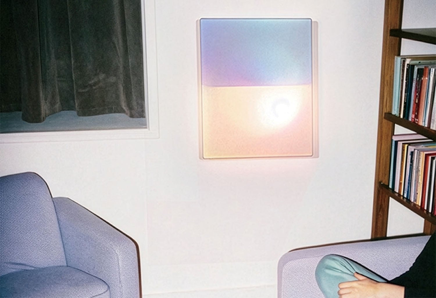 A pastel-colored light panel glows on a white wall between a blue armchair and a bookshelf; part of a person's arm and leg are visible in the foreground.