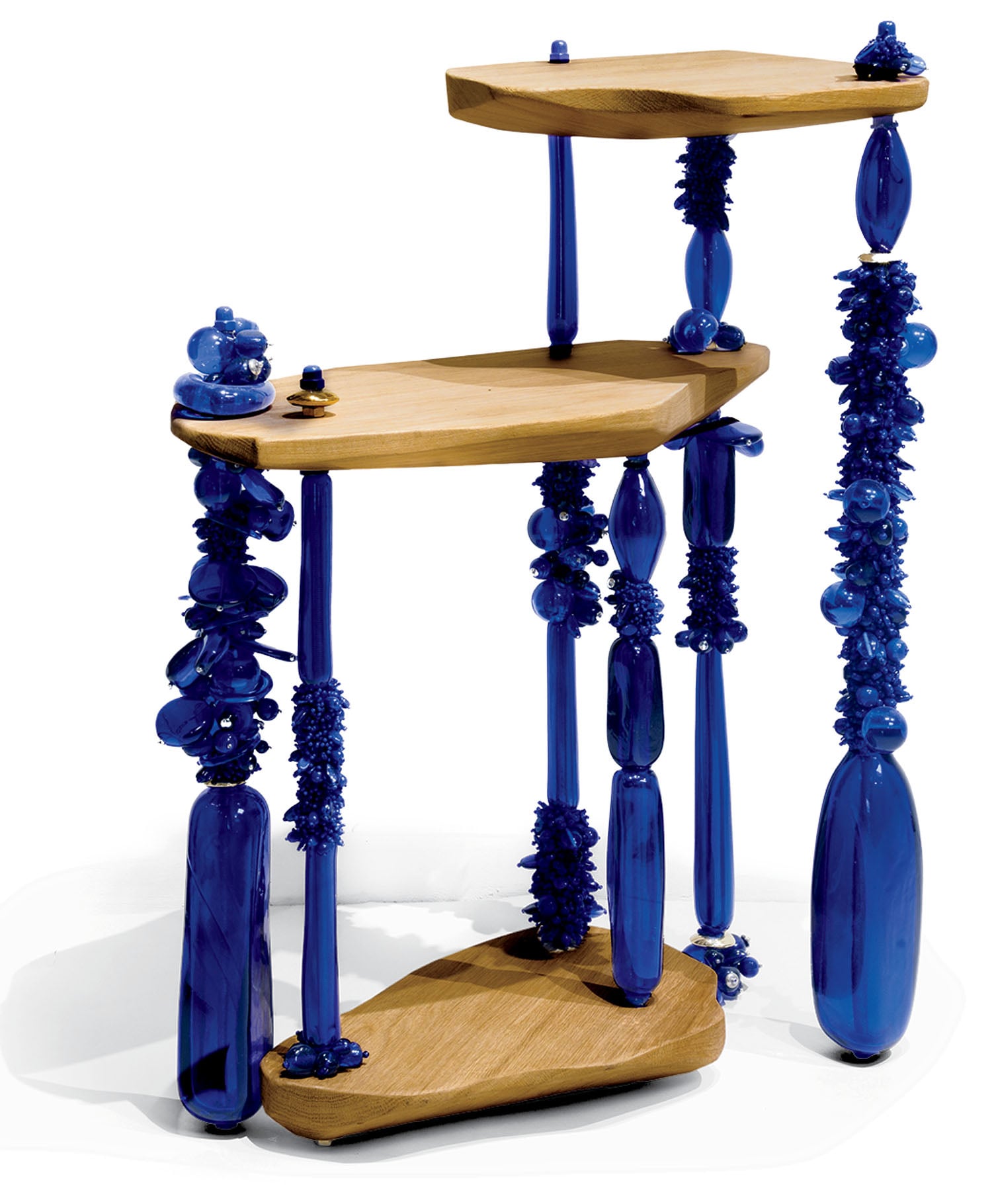 Three wooden shelves are supported by sculptural blue glass legs adorned with various textured and rounded glass details.