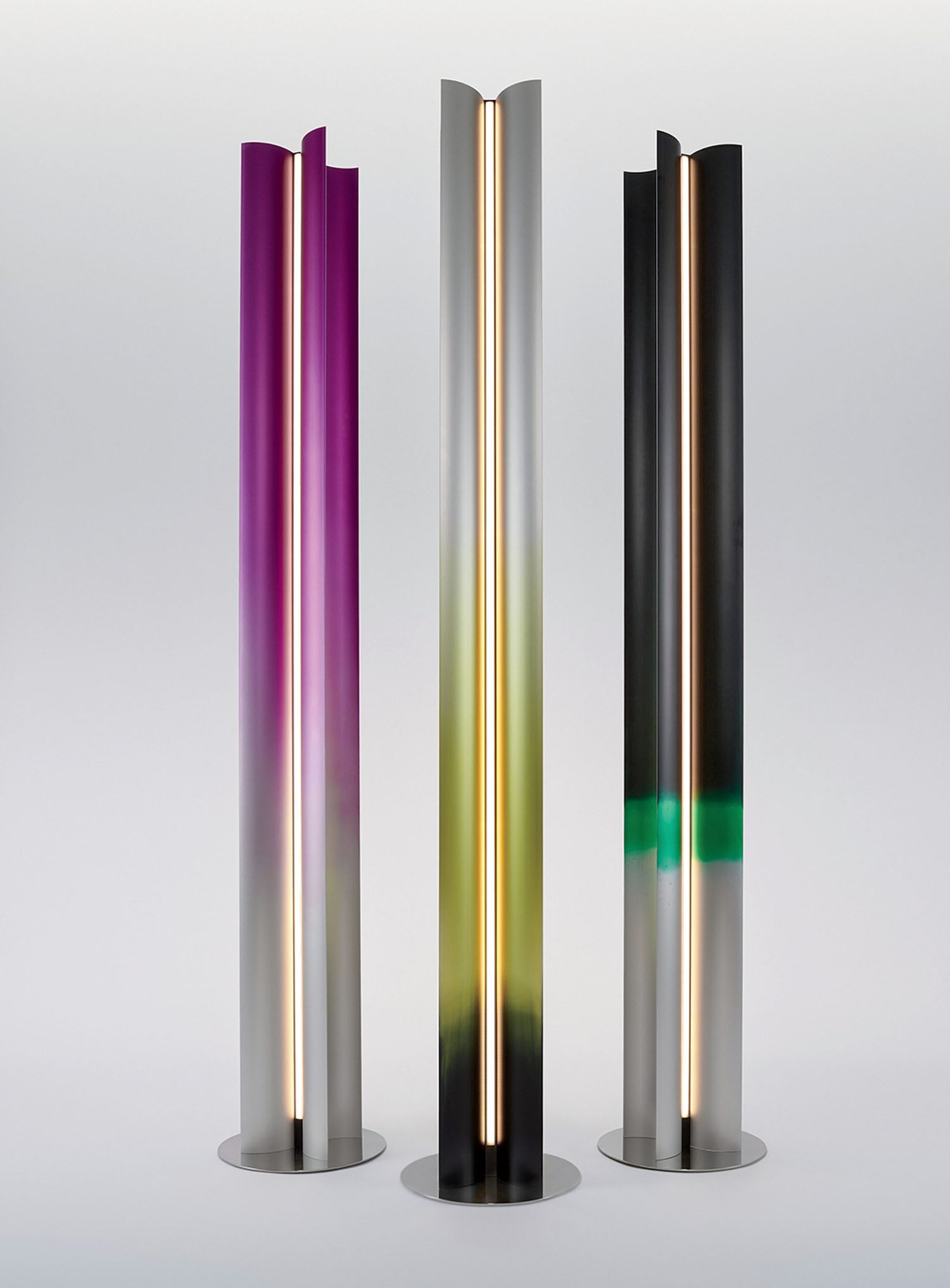 Three tall, cylindrical floor lamps with vertical LED lights stand side by side; each has a gradient finish in purple, yellow-green, and black-green tones.