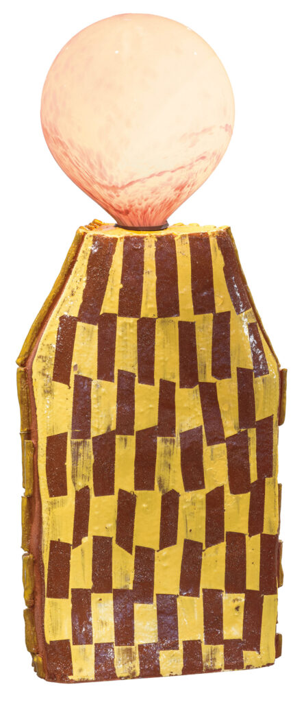 A geometric ceramic sculpture with a yellow and brown checkered body, topped with a large pinkish glass sphere.