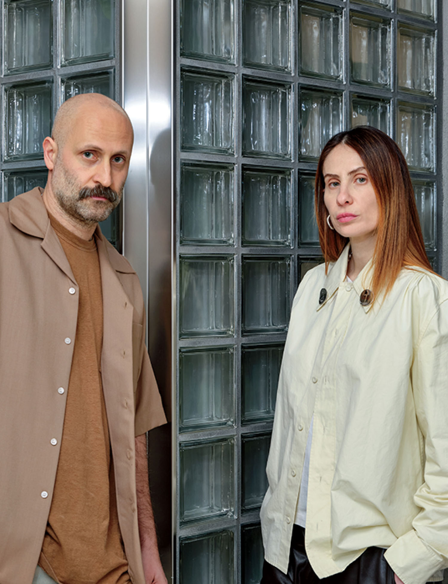 Two adults standing side by side in front of a wall made of glass blocks, looking at the camera with neutral expressions.