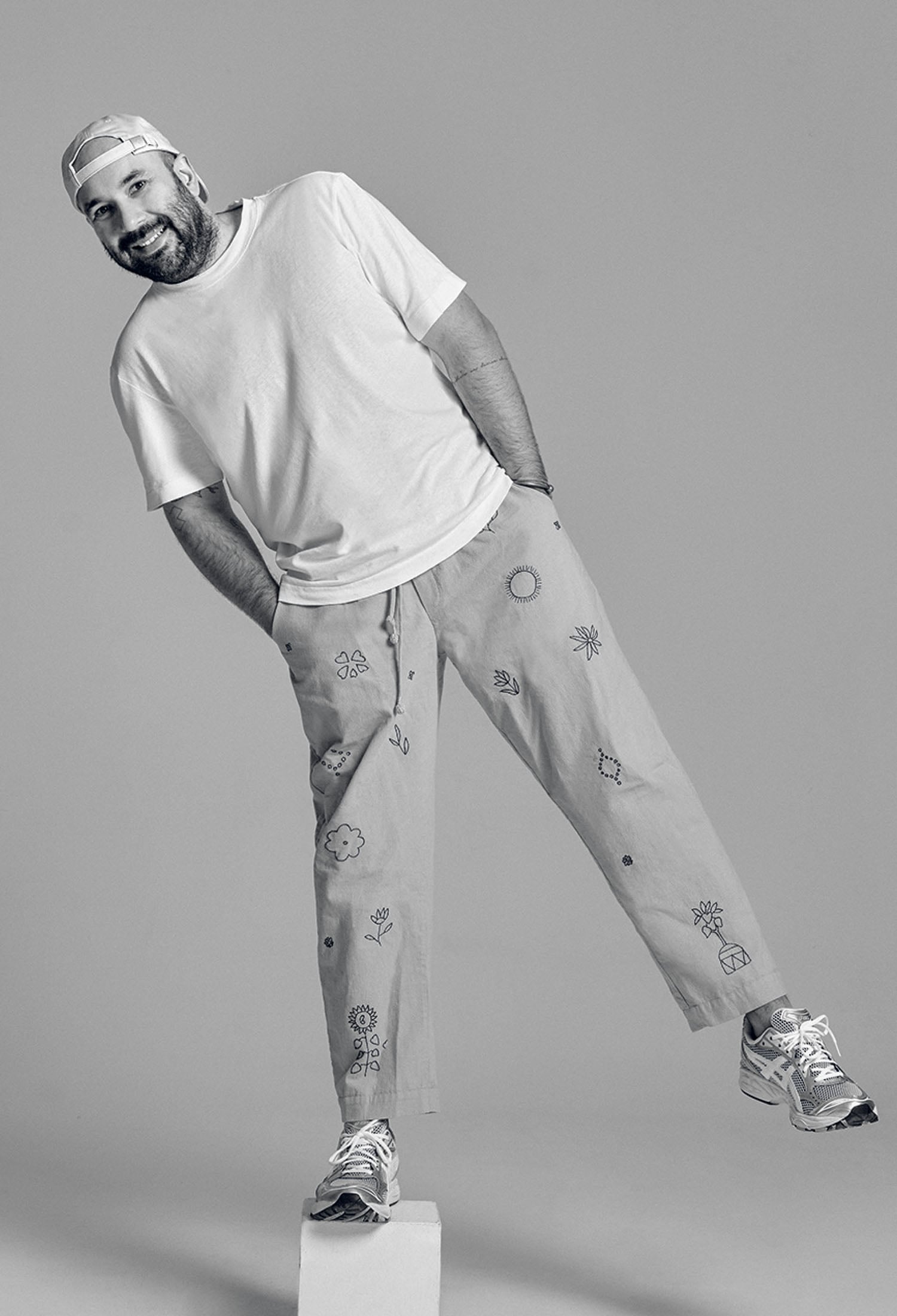 A man in a white t-shirt, patterned pants, sneakers, and a cap leans sideways with hands in pockets, standing on a white block against a plain background.