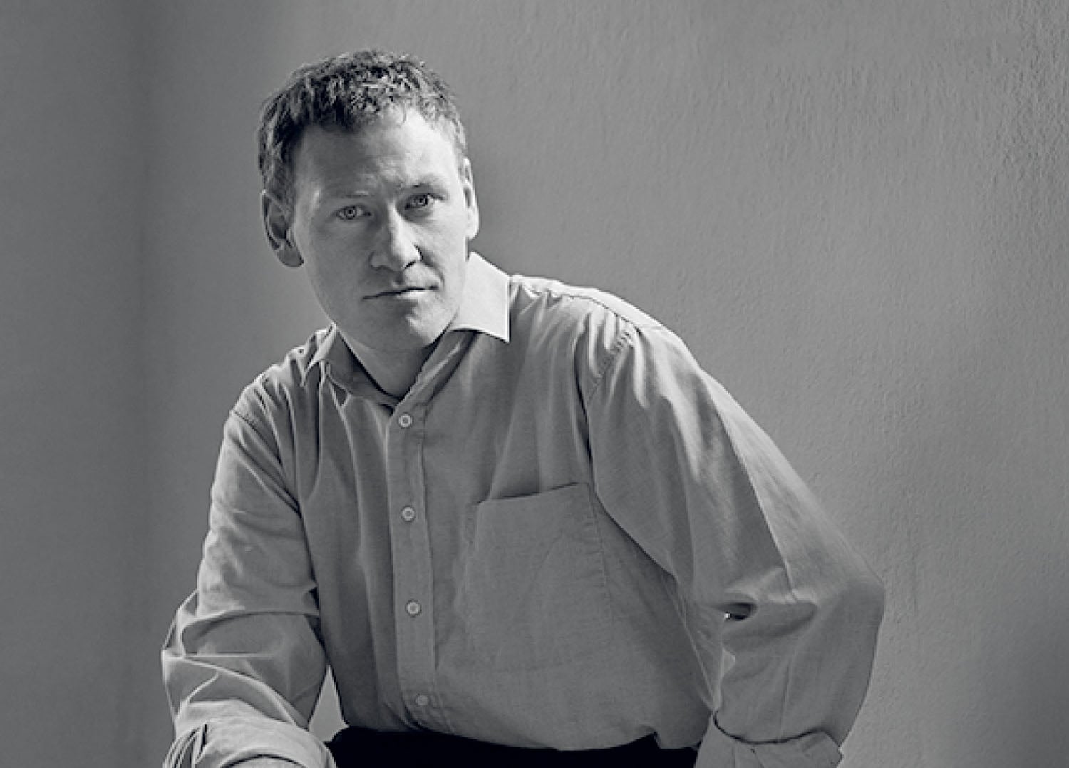 A man with short hair wearing a button-down shirt sits against a plain wall, looking slightly to the side with a neutral expression.