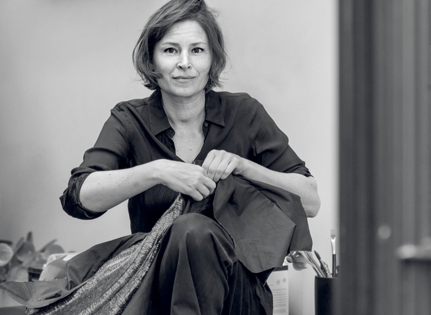 A woman sits indoors, holding fabric in her hands and looking toward the camera. The image is in black and white.