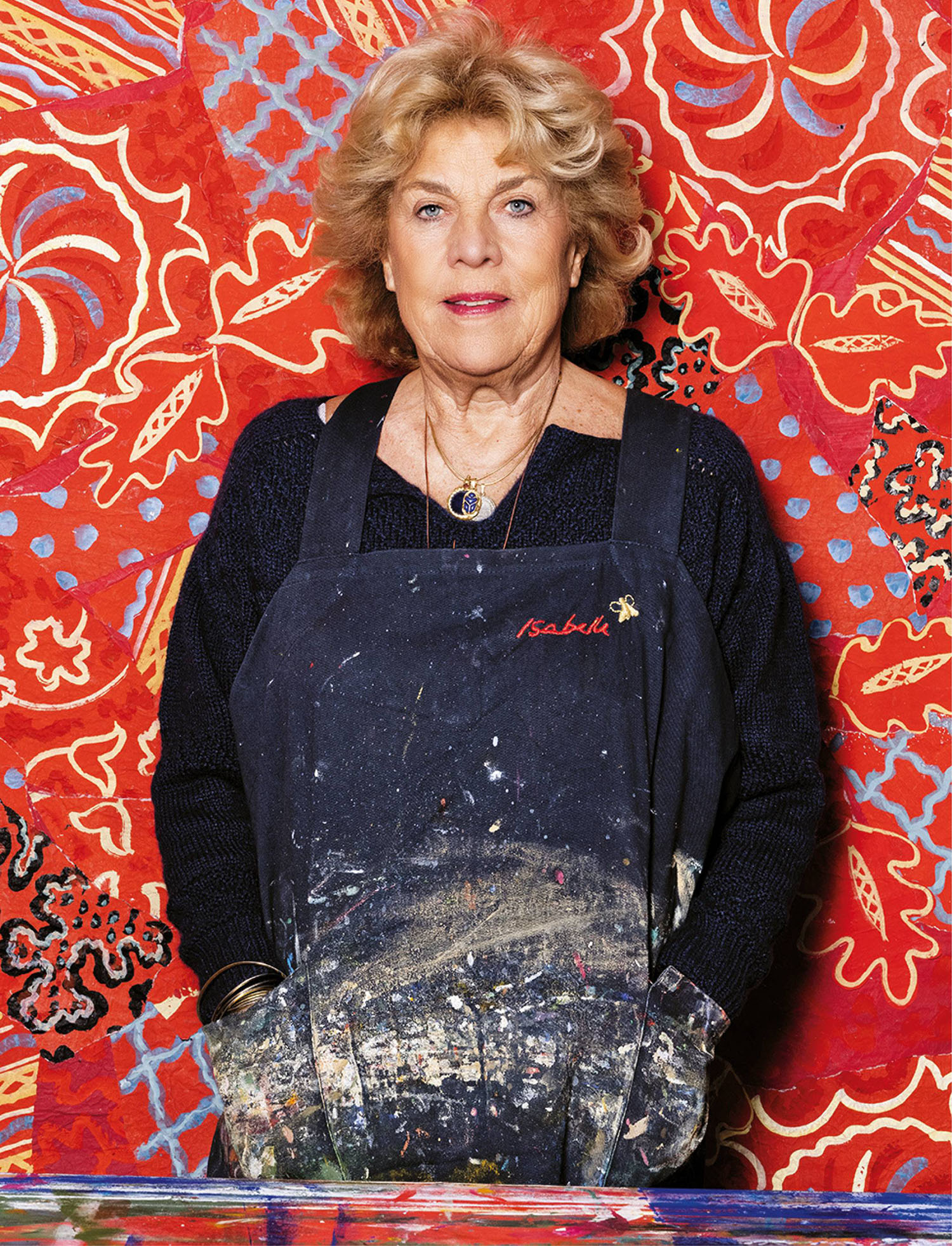 A woman wearing a paint-stained apron stands in front of a vibrant, patterned red backdrop with floral and abstract designs.