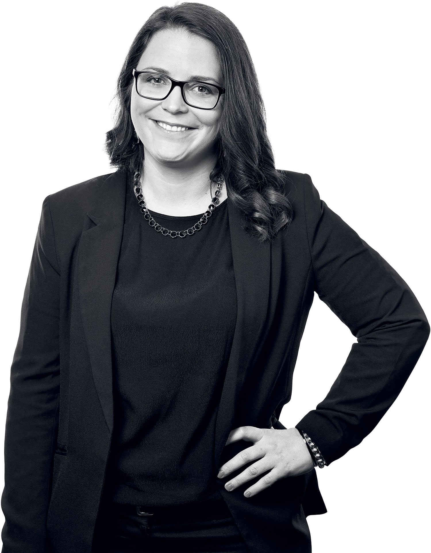 Woman wearing glasses and a dark business suit stands with one hand on her hip, smiling at the camera against a white background.