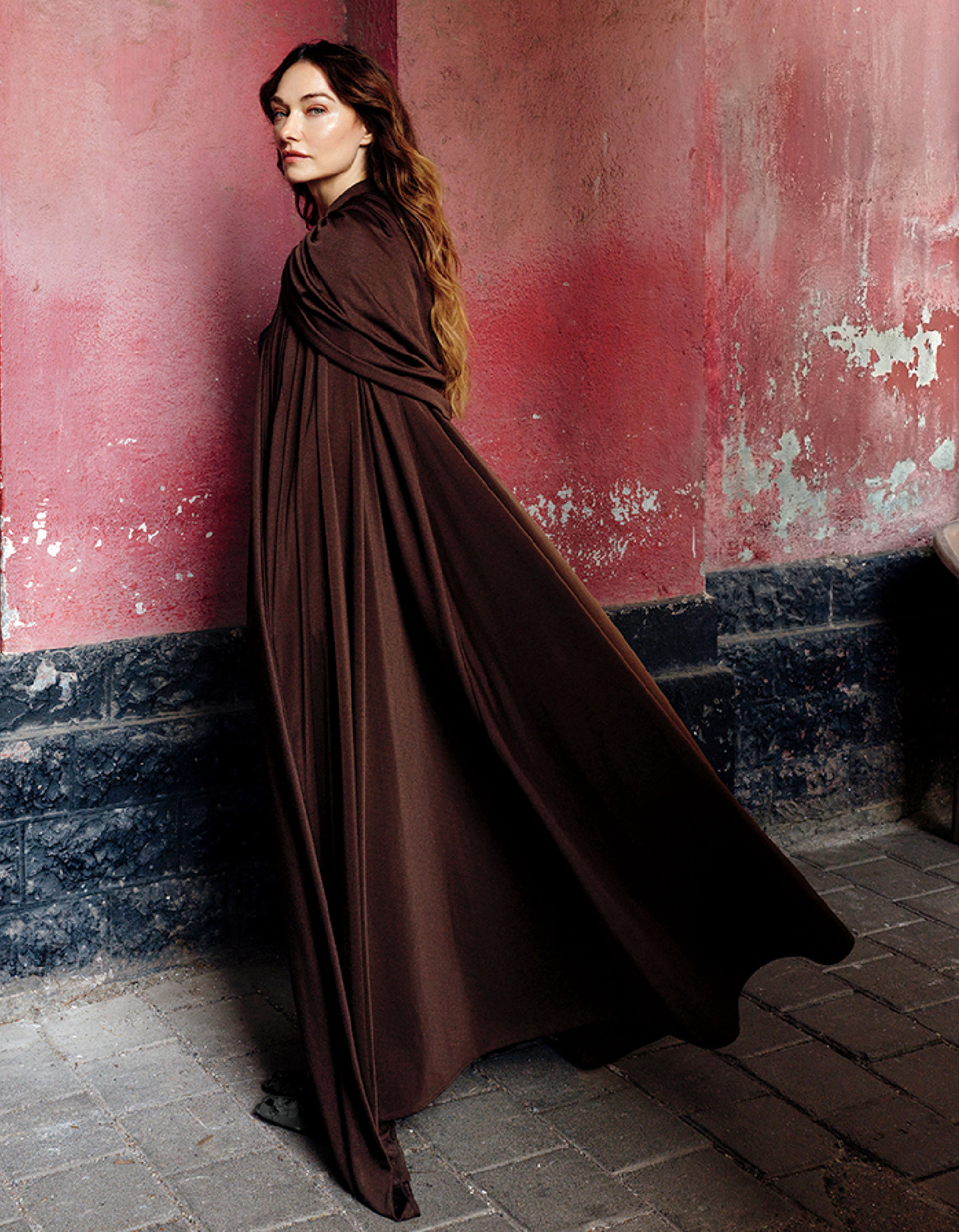 Person with long hair wearing a flowing brown cloak stands against a weathered pink and black wall on a stone-paved surface.