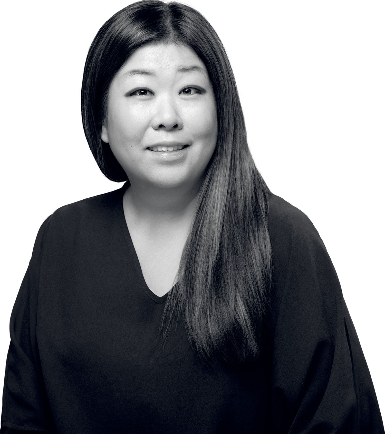 A woman with long straight hair, wearing a dark V-neck top, looks slightly to the side and smiles in a black and white portrait against a plain background.