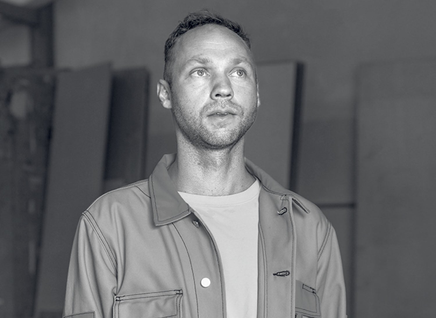 A man in a light-colored jacket and t-shirt stands indoors, looking slightly upward. The image is in black and white.
