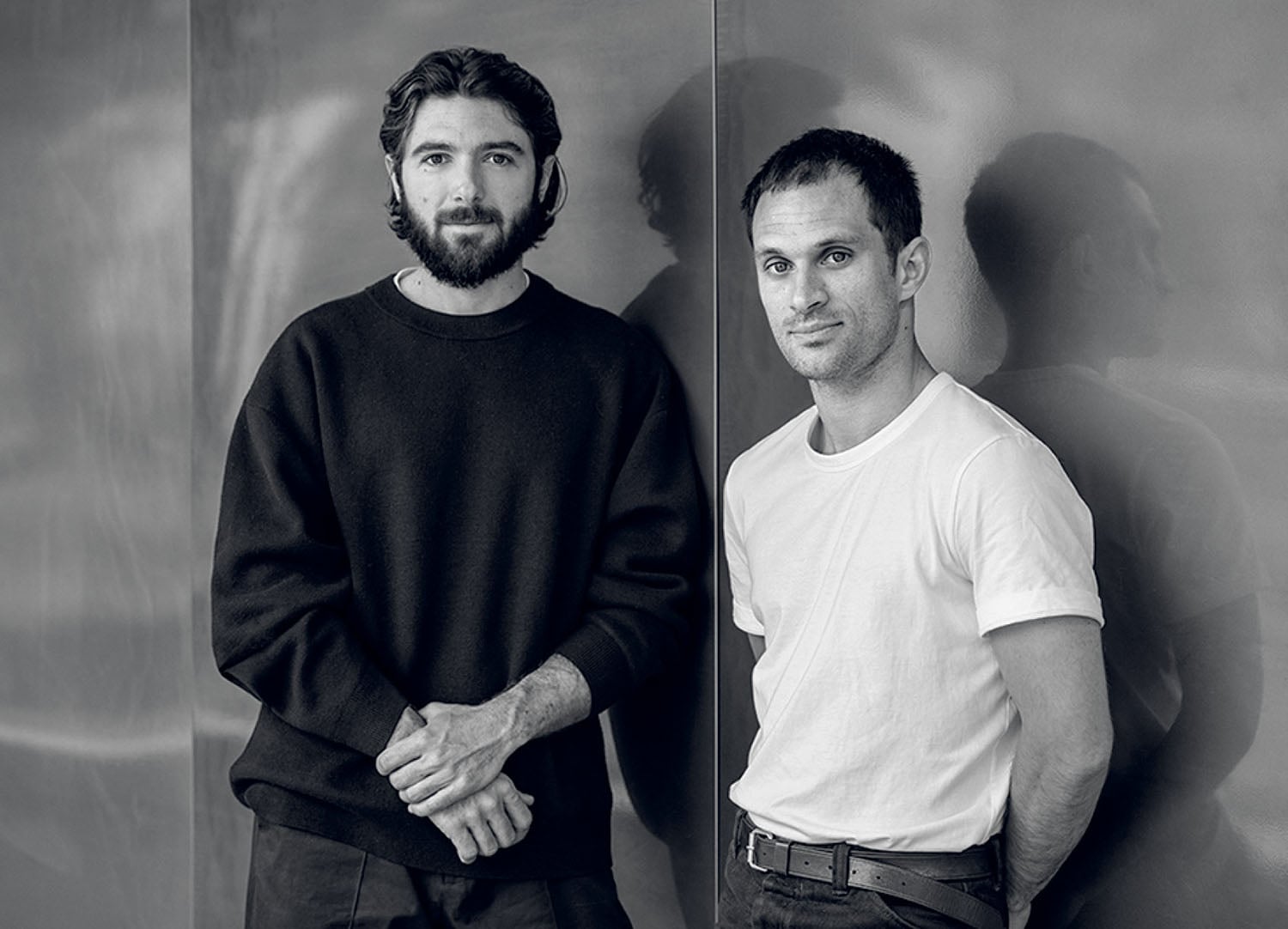 Two men stand side by side against a reflective wall; one wears a dark sweater and has a beard, the other wears a white t-shirt and stands with his hands behind his back.