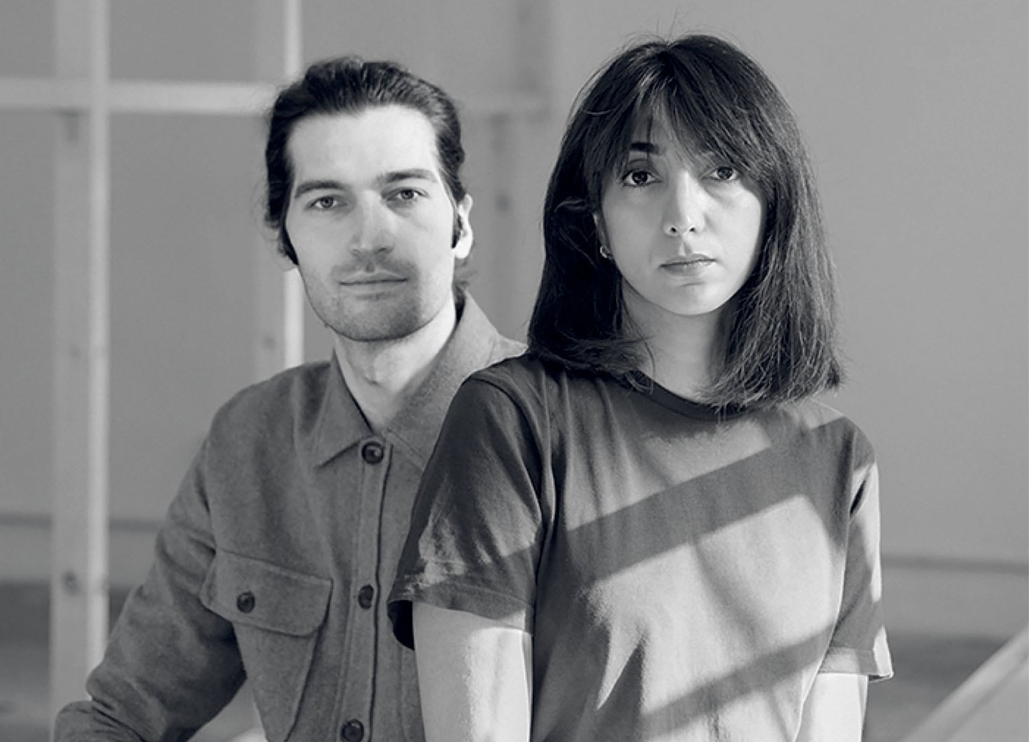 Two people sit indoors; one with long dark hair and a buttoned shirt, the other with medium-length hair and a T-shirt, both looking at the camera in natural lighting.
