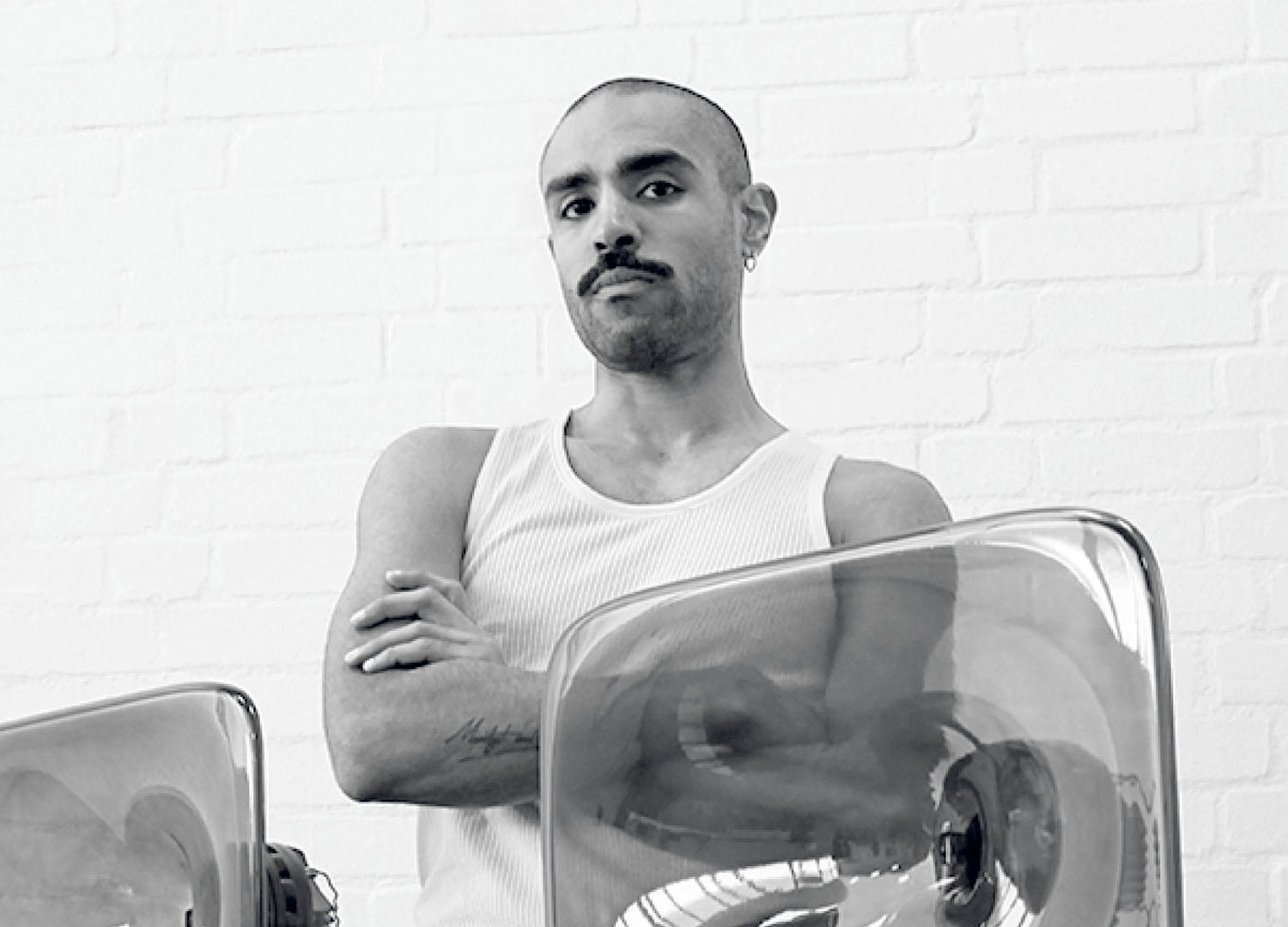 A person with a shaved head and mustache stands with arms crossed, wearing a white tank top, in front of a white brick wall and transparent objects.