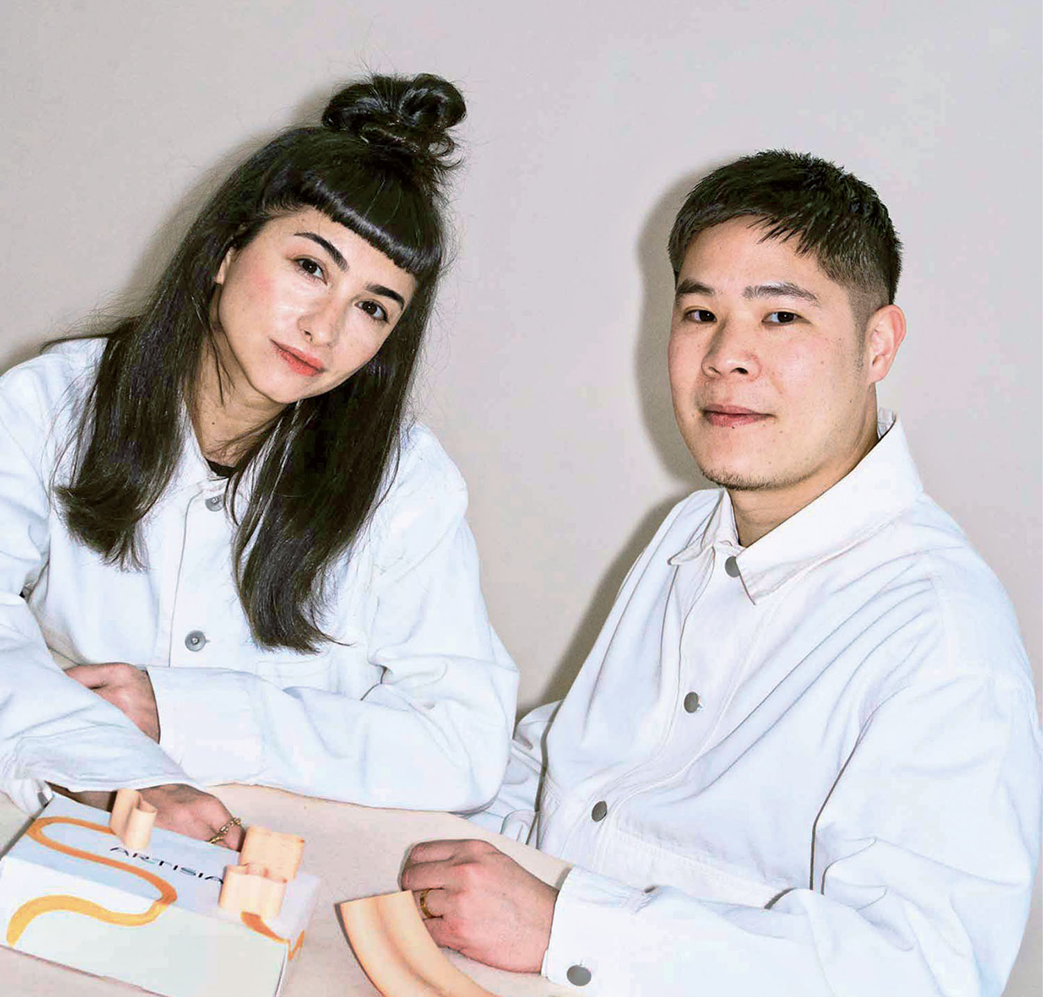 Two people wearing white shirts sit at a table with pastel-colored objects in front of them, against a plain background.
