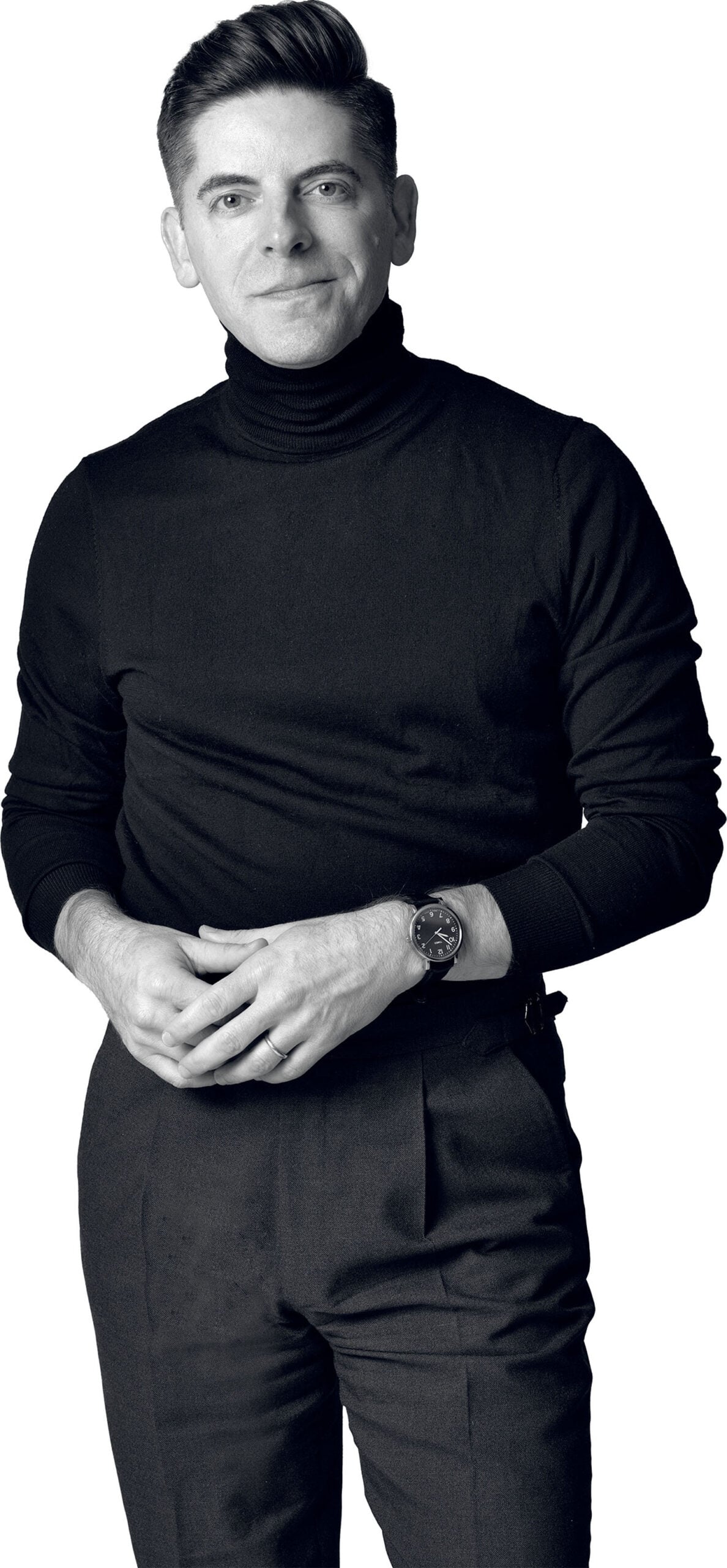 Person standing and facing forward, wearing a black turtleneck sweater, dark pants, and a wristwatch, with hands loosely clasped.