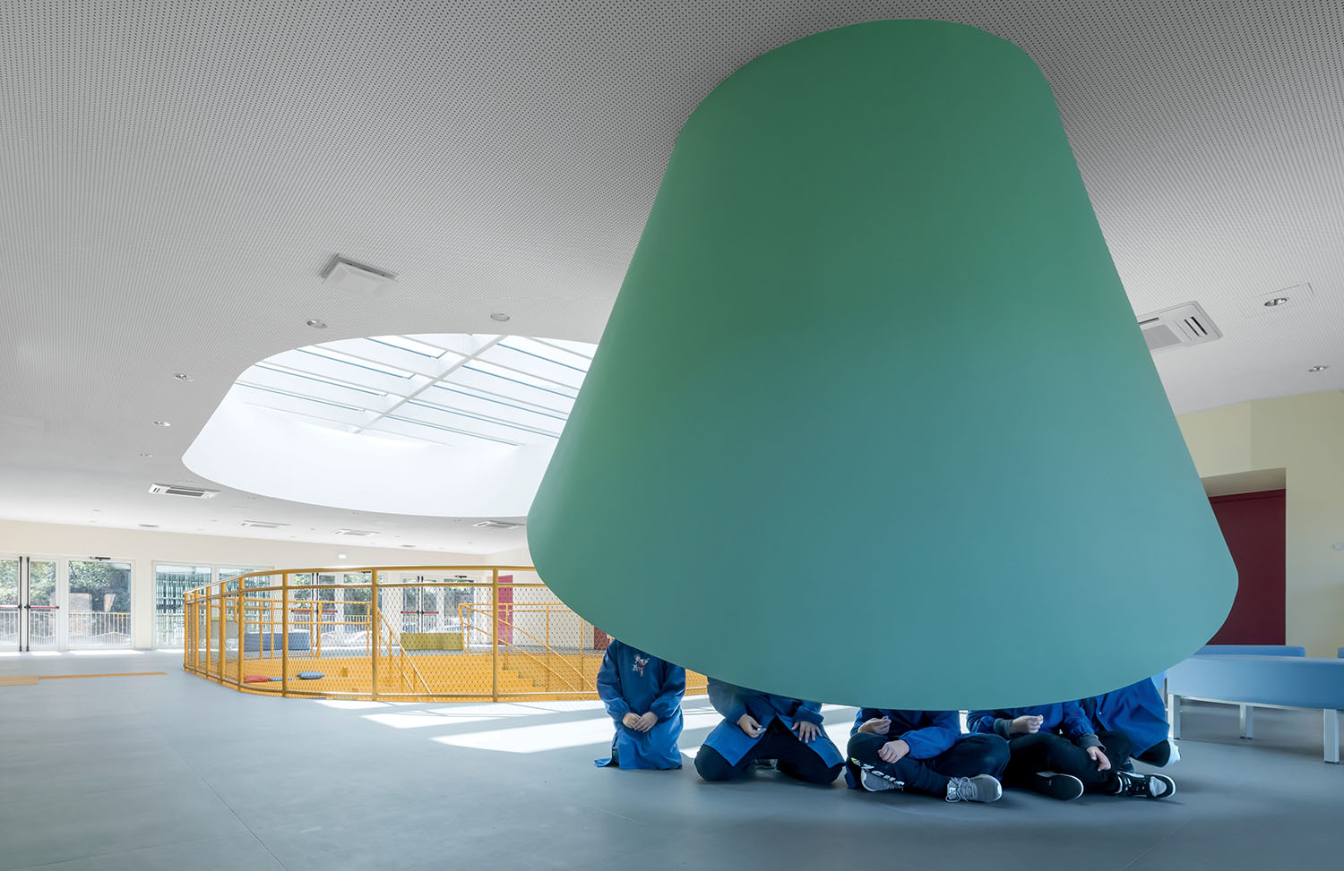 Six people in blue uniforms sit beneath a large, green, cone-shaped structure in a modern, spacious room with skylights and a yellow fenced area.