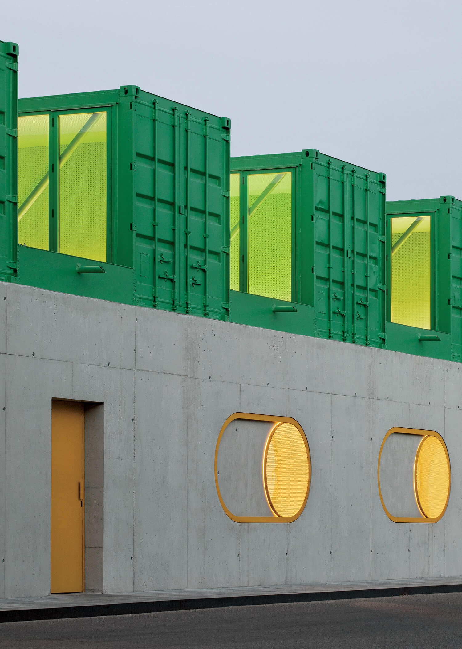 A modern building with a concrete base featuring yellow-framed round windows and door, topped with green shipping containers with yellow-lit interiors.