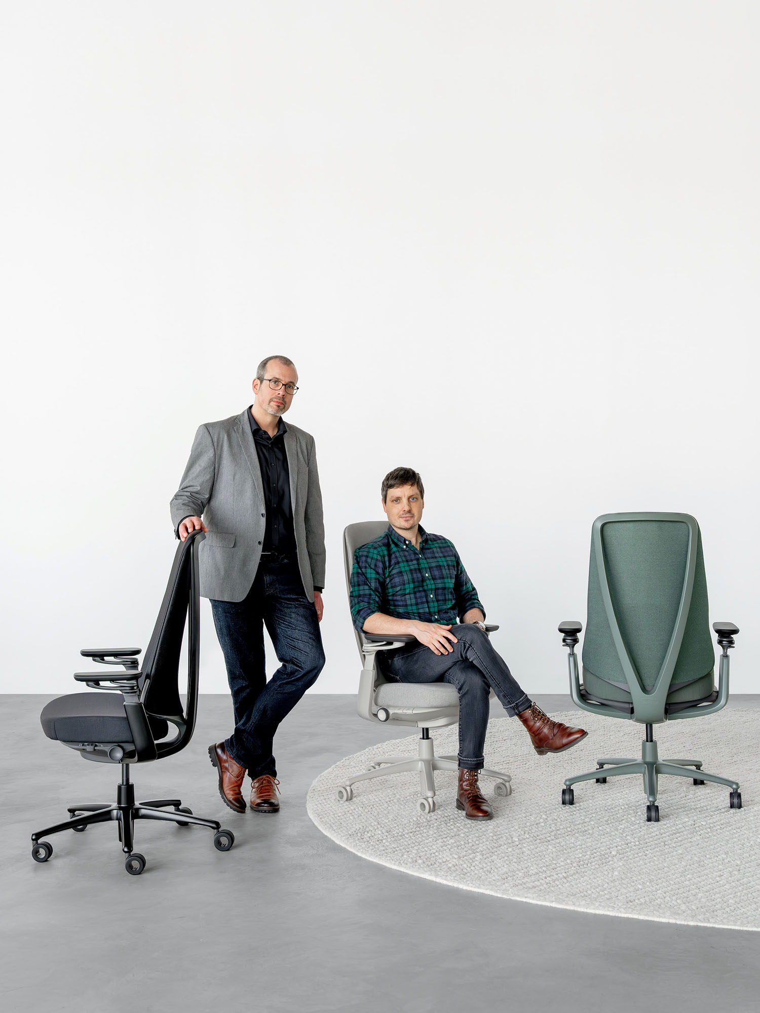 Two men pose with three office chairs in a minimalist room; one stands next to a black chair while the other sits on a gray chair, and a green chair is also displayed.