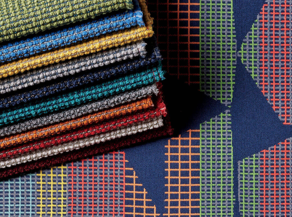 A stack of textured fabric swatches in various colors sits on top of a dark blue fabric with a multicolored geometric grid pattern.