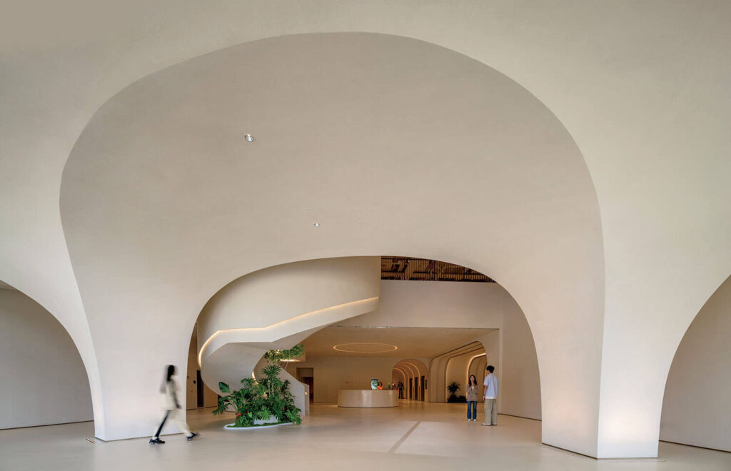 Spacious modern lobby with curved archways, a spiral staircase with greenery, and three people near the reception desk.