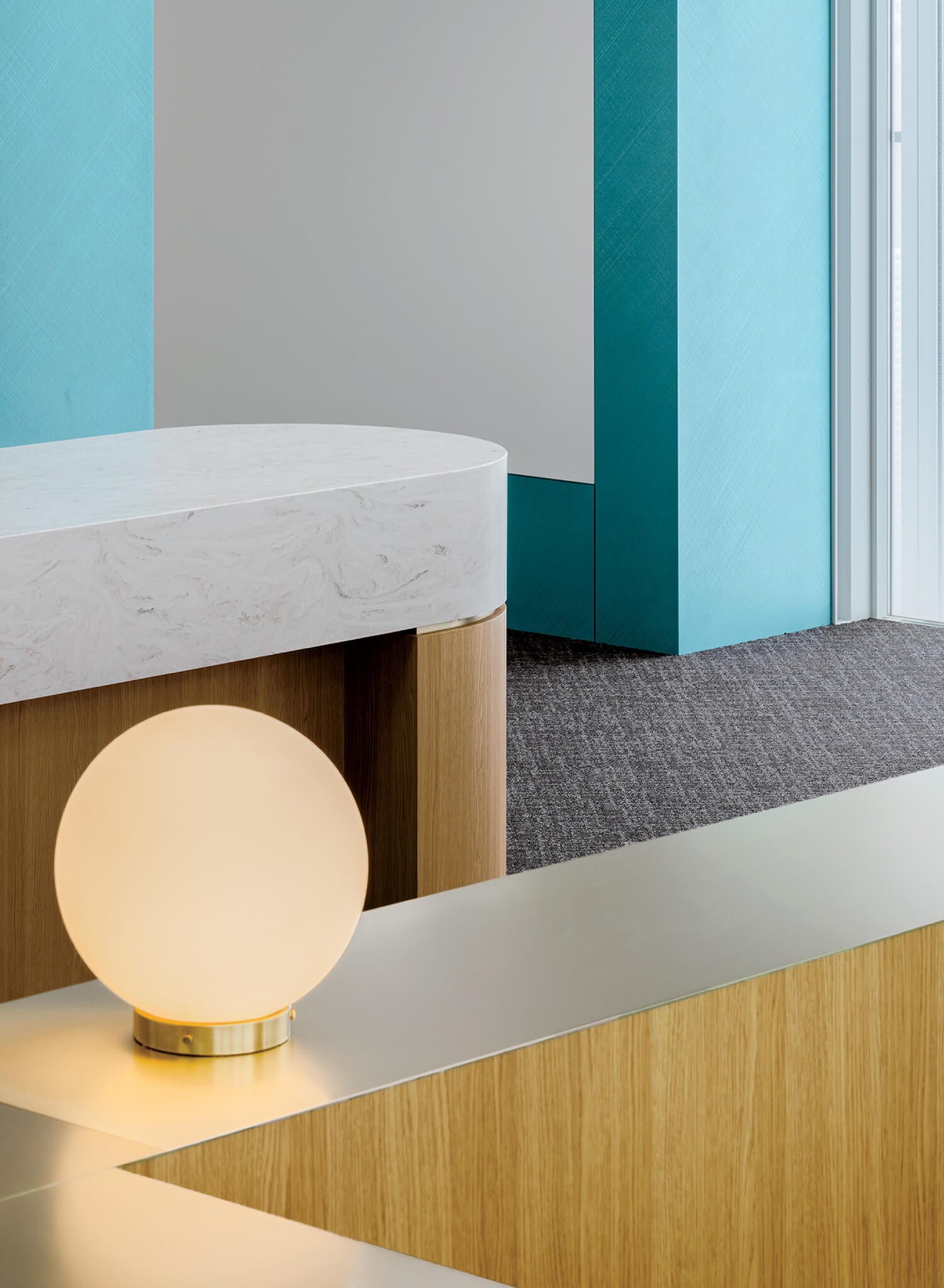A round white lamp with a gold base sits on a light wood and metal surface, with turquoise and gray walls and textured gray carpet in the background.
