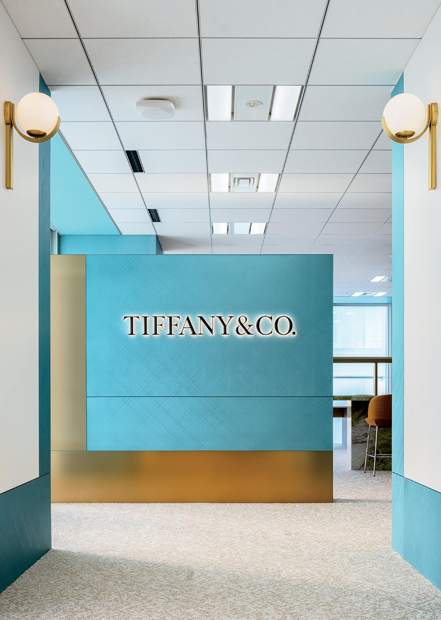 A modern office interior features a blue wall with the illuminated Tiffany & Co. logo, gold accents, and contemporary wall sconces.