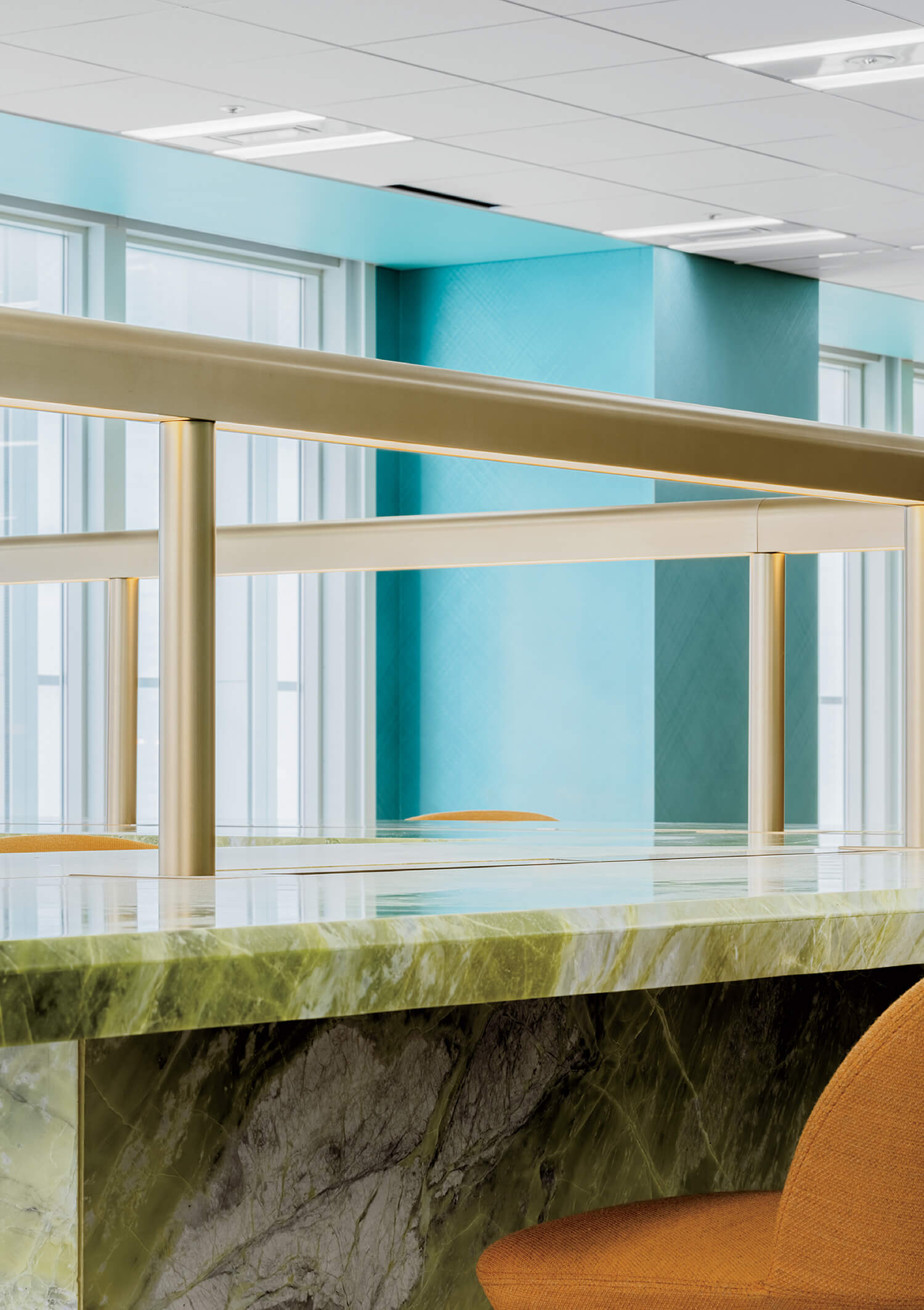 A modern interior space featuring a green marble countertop, brass railing, turquoise walls, large windows, and an orange upholstered chair.