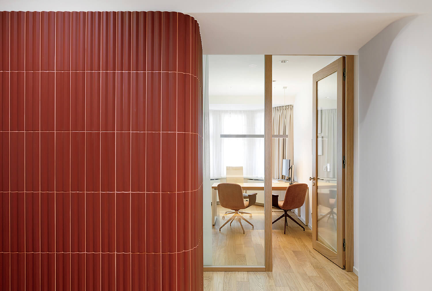 A modern office with wooden floors, glass doors, two chairs, a desk, and a curved red tiled wall on the left.