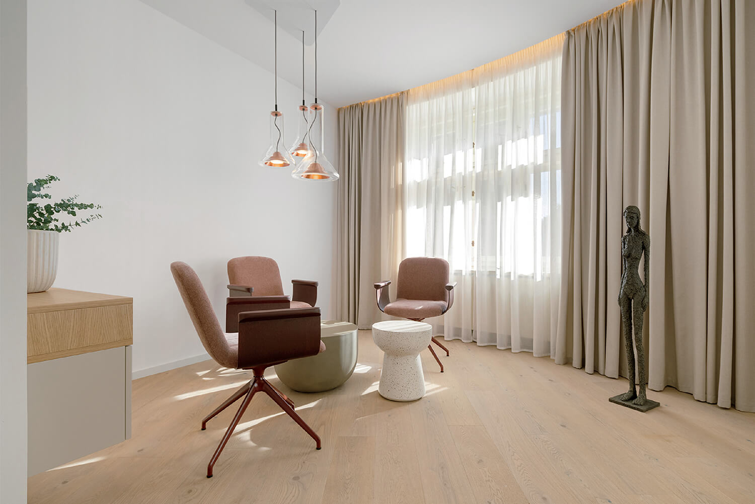Minimalist room with two pink armchairs, a small white table, pendant lights, a potted plant, a standing sculpture, and large beige curtains covering a window.