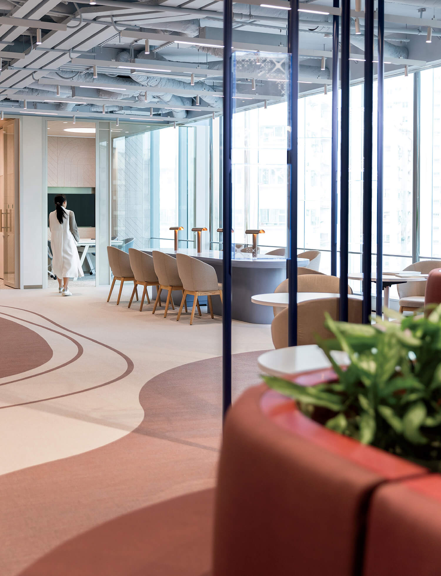 Modern office lounge with contemporary furniture, large windows, and a person walking in the background; green plants visible in the foreground.
