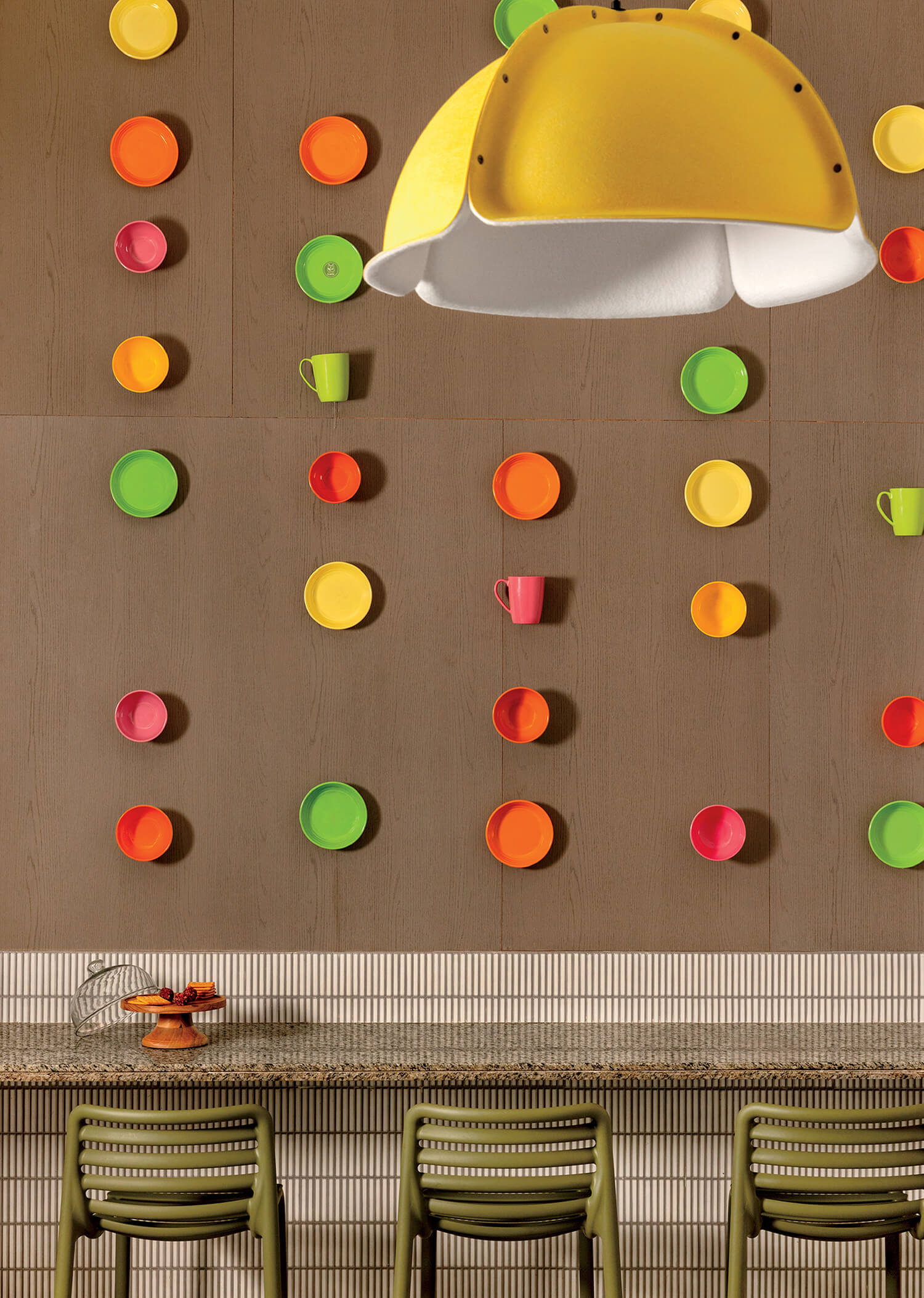 A wall decorated with red, orange, yellow, and green cups and plates arranged in vertical lines above a counter with three stools and a large yellow pendant lamp overhead.