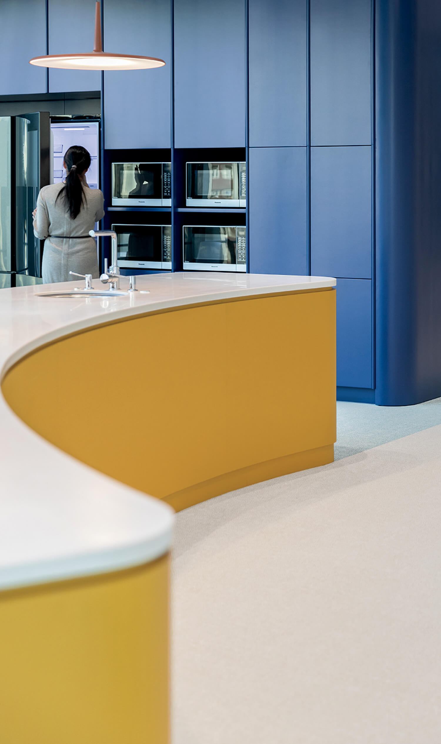 A person stands at an open refrigerator in a modern kitchen with blue cabinets, built-in microwaves, and a curved yellow island.