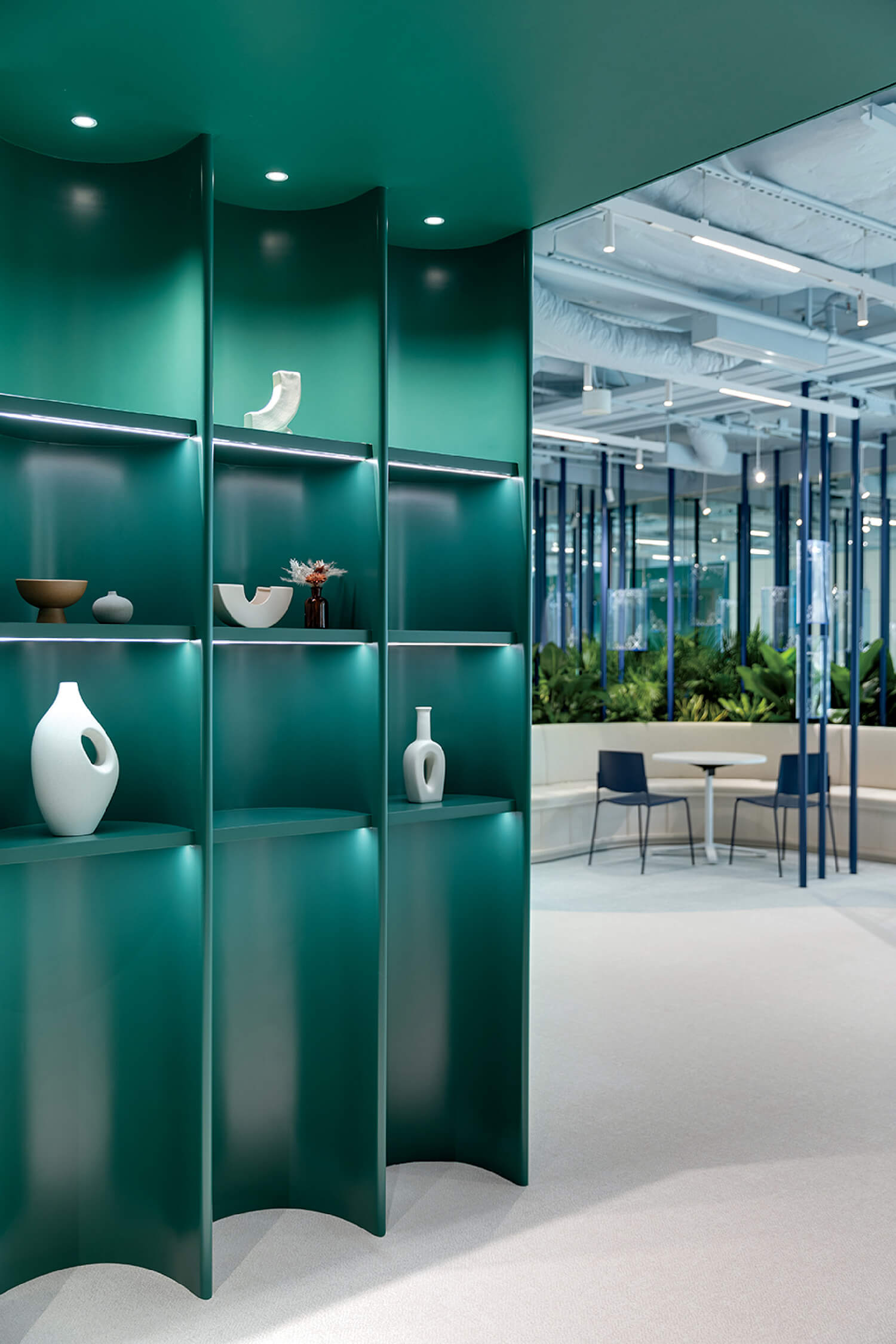 Curved green shelving displays minimalist ceramic vases and bowls; modern office space with plants and seating is visible in the background.