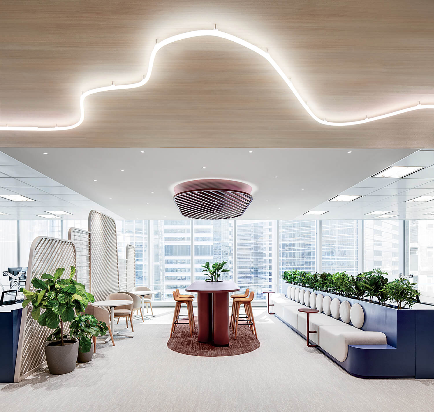 Modern office space with plants, a long cushioned bench, tables and chairs, and large windows providing natural light. Curved ceiling light and cityscape visible through the windows.