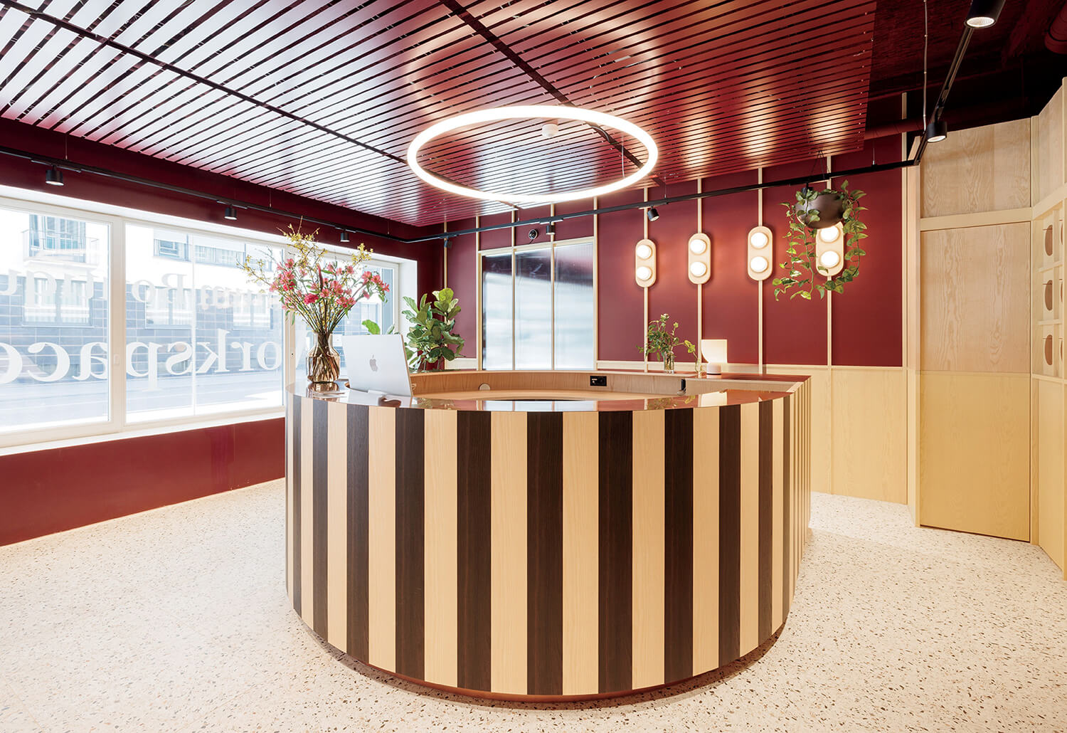 Modern reception area with a striped circular desk, hanging lights, plants, and large windows, featuring a red ceiling with a circular light fixture.