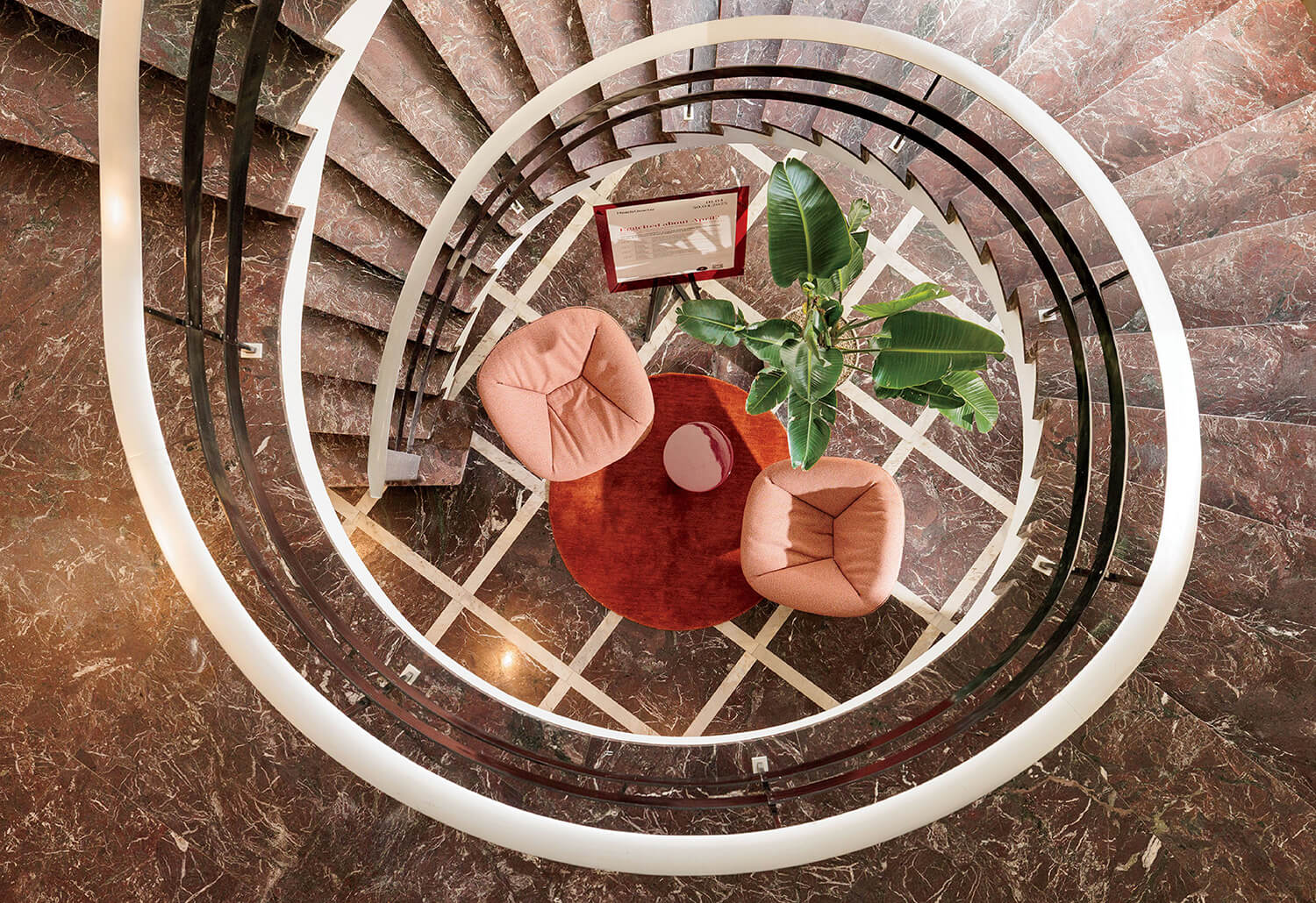 Top-down view of a spiral staircase surrounding two pink chairs, a small round table, a red rug, and a green potted plant on a marble floor.