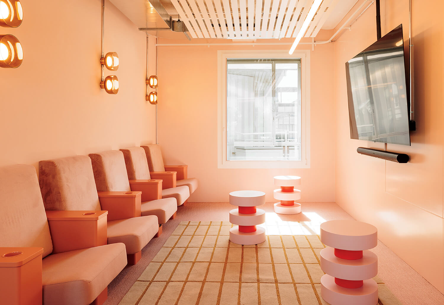 A modern lounge with peach-colored walls, cushioned chairs, round side tables, a grid-patterned rug, wall lights, a large window, and a wall-mounted TV.