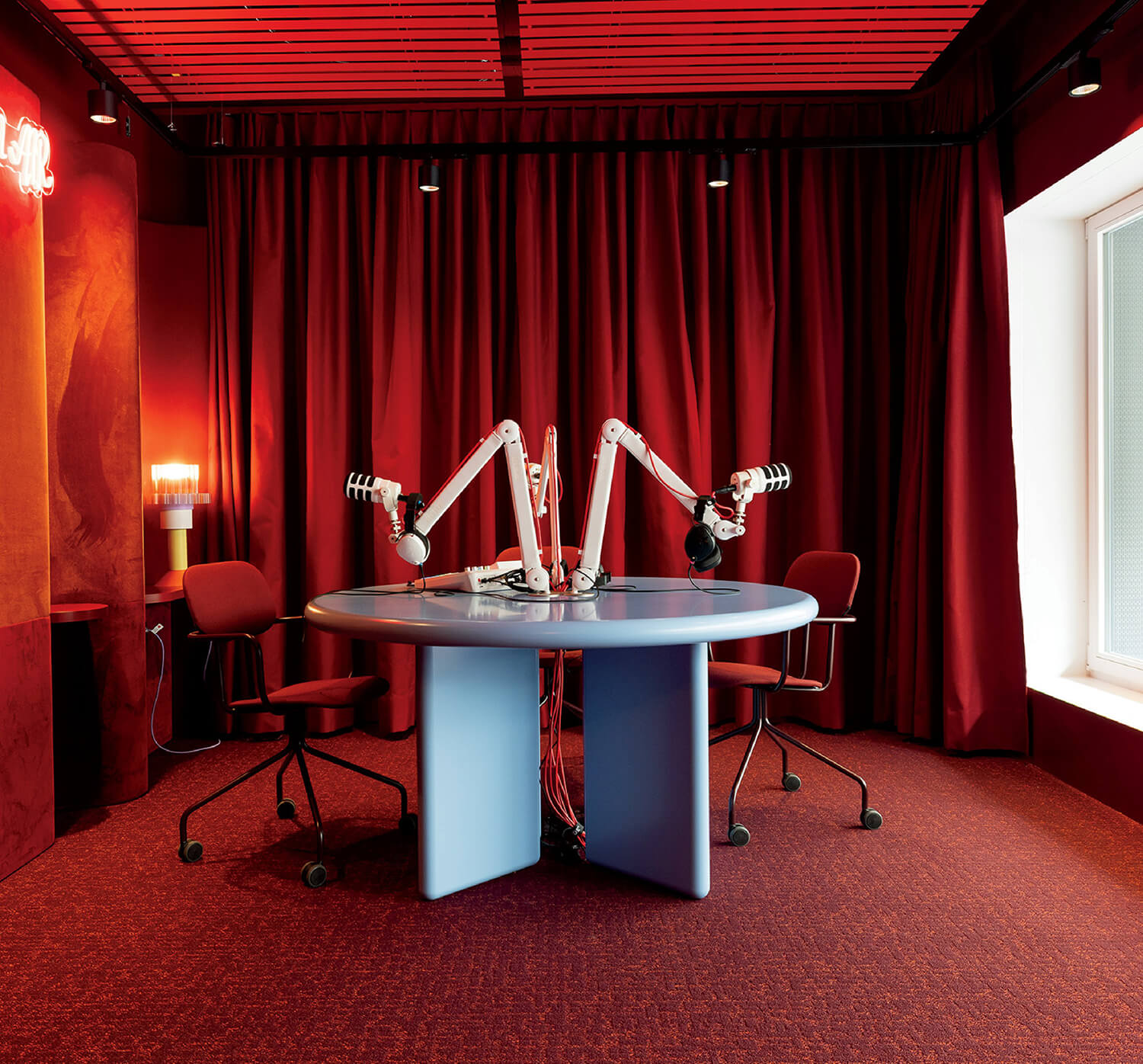 A modern podcast studio with two microphones on adjustable arms, red chairs, red curtains, and a round blue table under warm lighting.