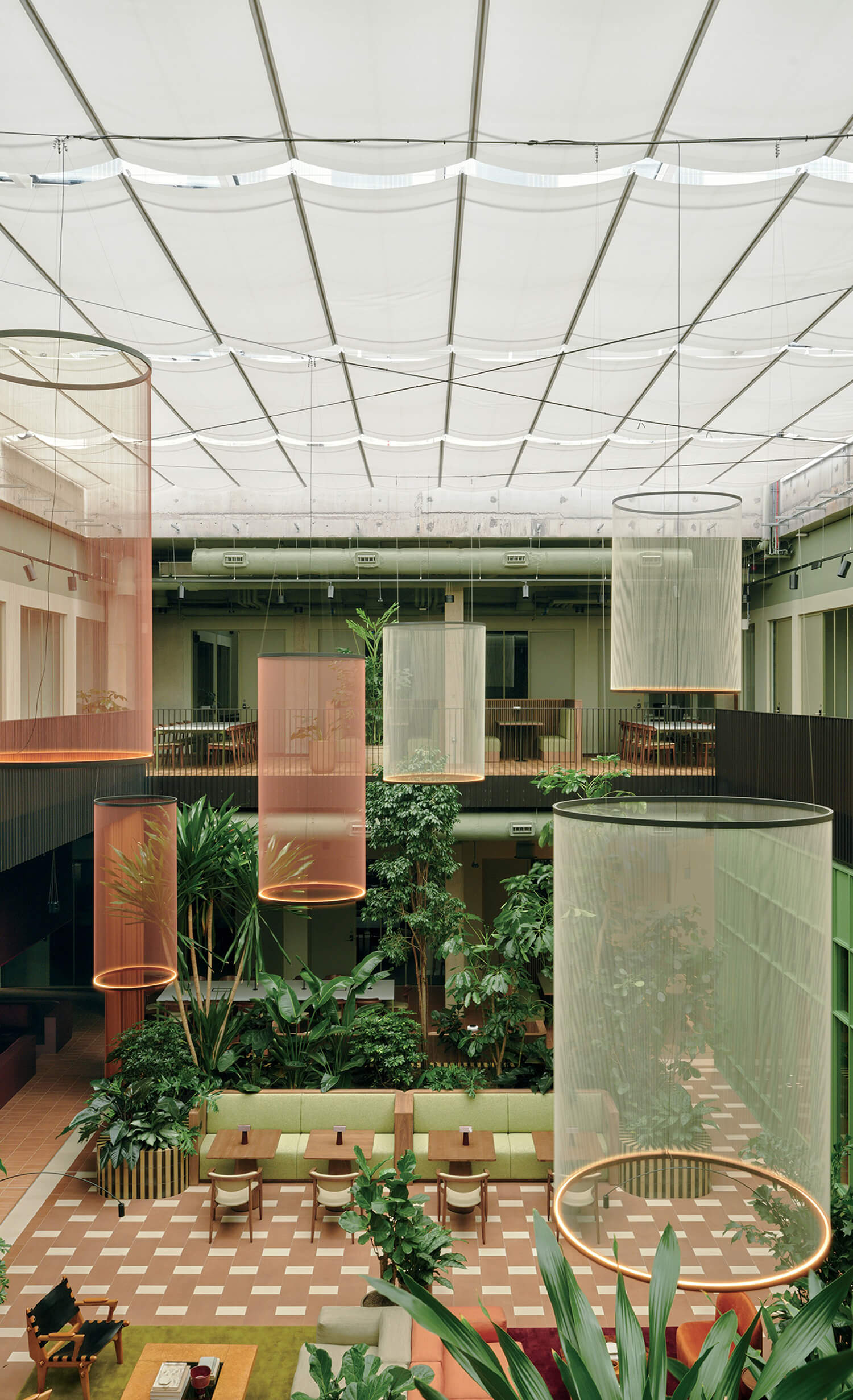 Modern indoor atrium with large hanging cylindrical mesh sculptures, lush green plants, checkerboard floor, and a mix of seating areas under a white translucent ceiling.