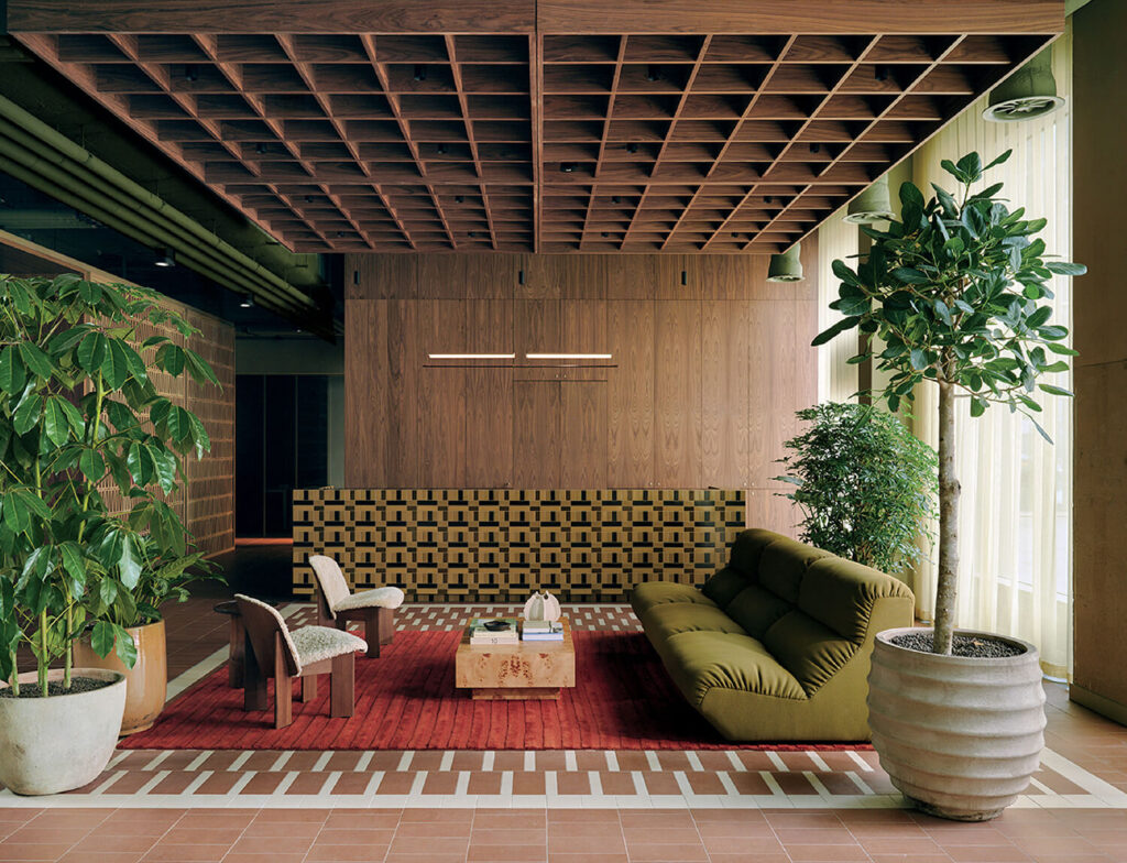 Modern lounge area with green sofa, patterned bench, wooden chairs, large potted plants, wooden ceiling grid, and a red rug on a tiled floor, lit by natural light from large windows.