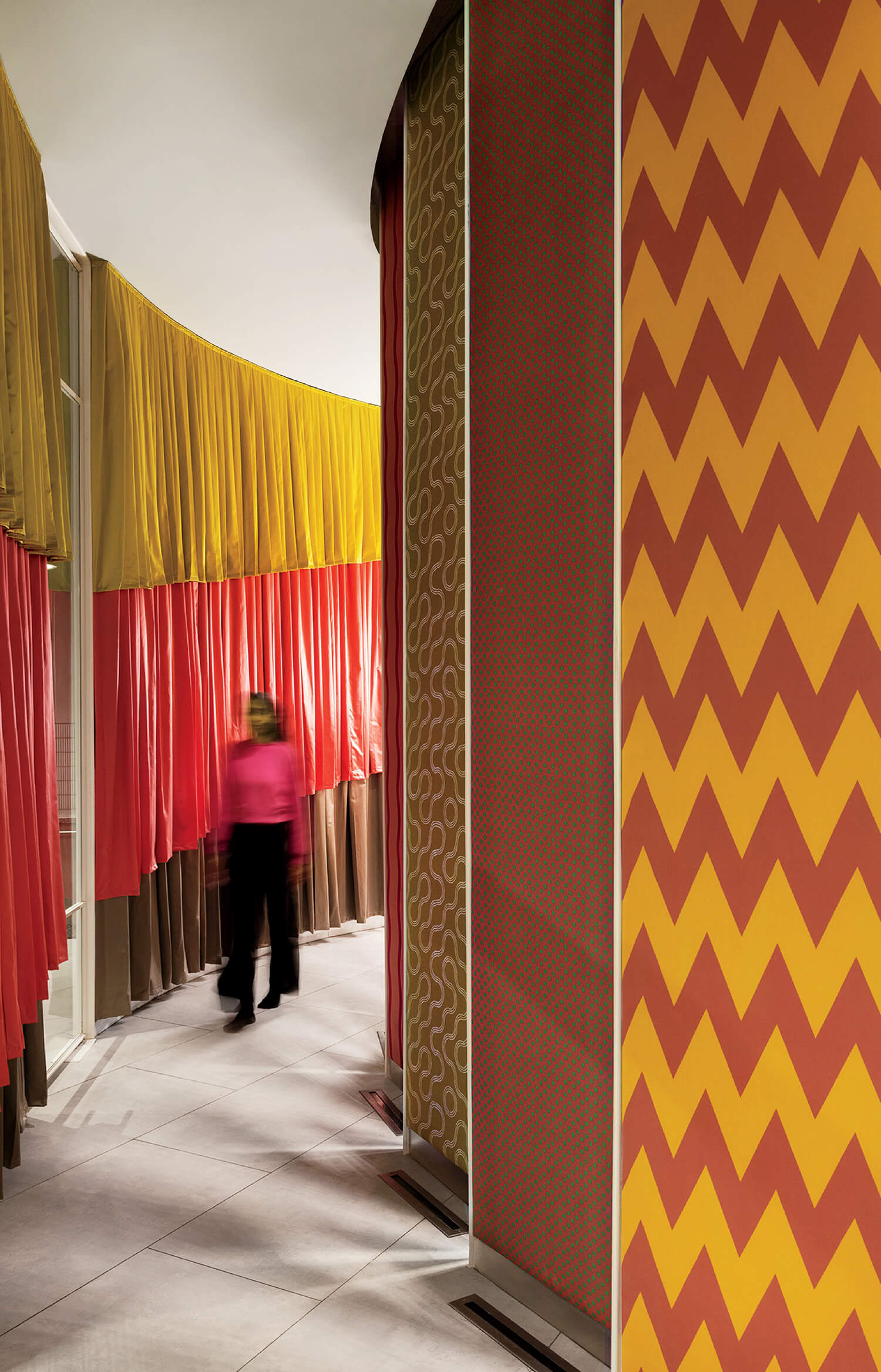 A person walks through a corridor with wavy yellow and red curtains and patterned walls featuring chevrons and abstract designs.
