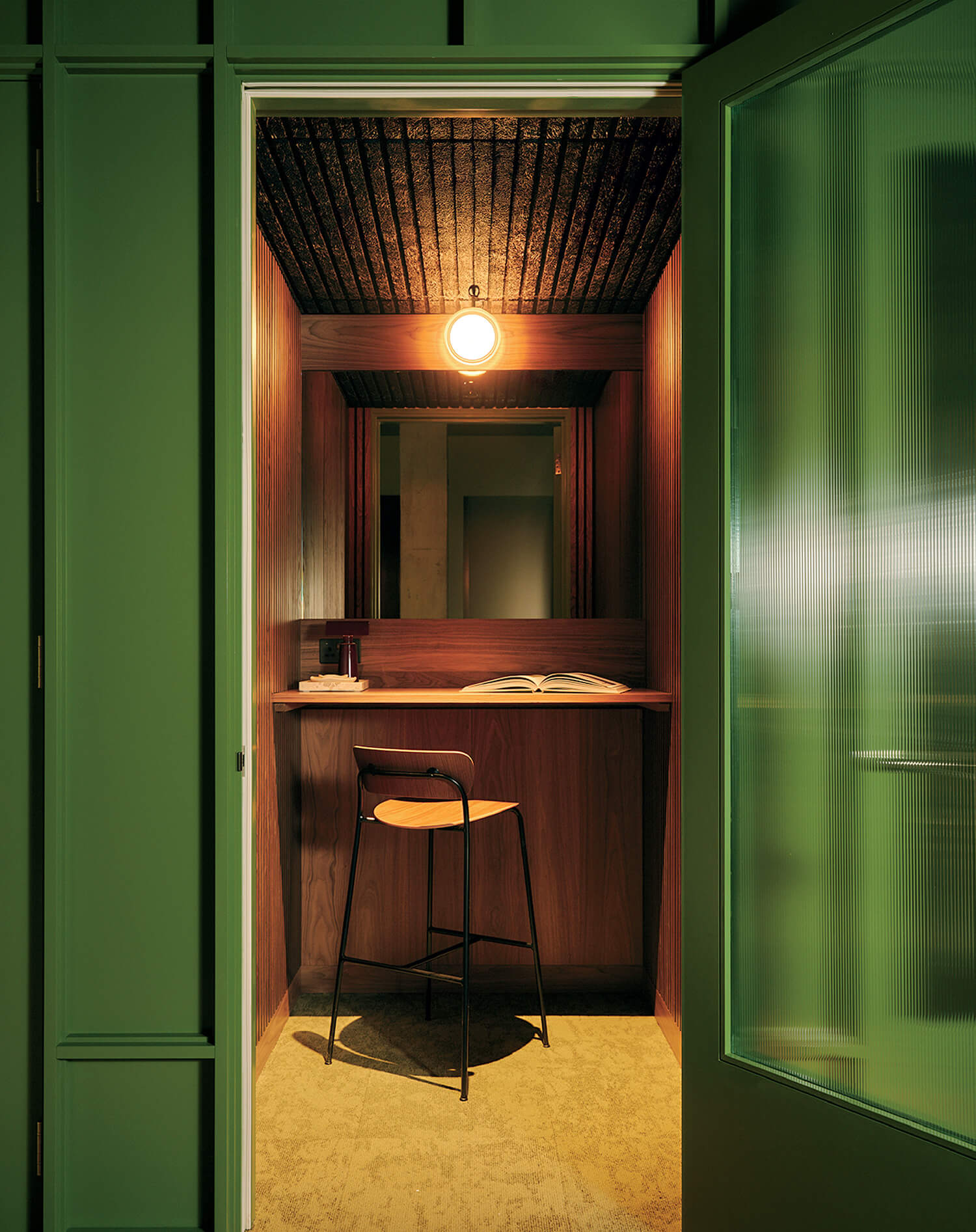 A small, wood-paneled room with a high counter, a single barstool, an open book, and a ceiling light, viewed through a green door.