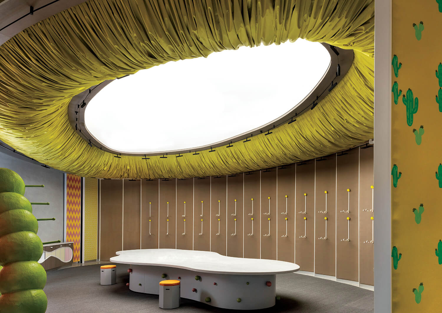 Modern room with large oval skylight draped in yellow fabric, curved white table, colorful stools, and lockers with yellow knobs. Green cactus decorations on the right wall.