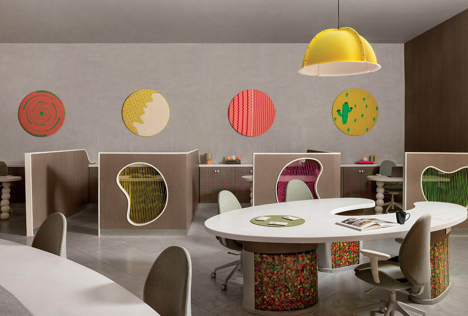 Modern office space with curved desks, individual cubicles, artistic wall decor, contemporary chairs, and a large yellow pendant lamp.