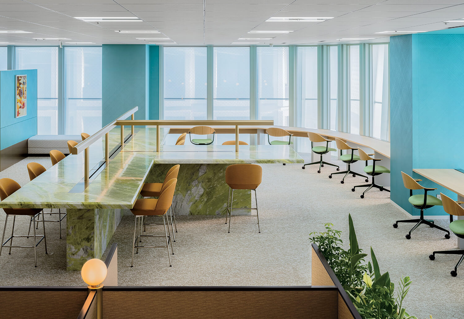 A modern office space with green marble communal tables, tan chairs, and large windows allowing natural light. Green plants and blue accent walls add color to the room.