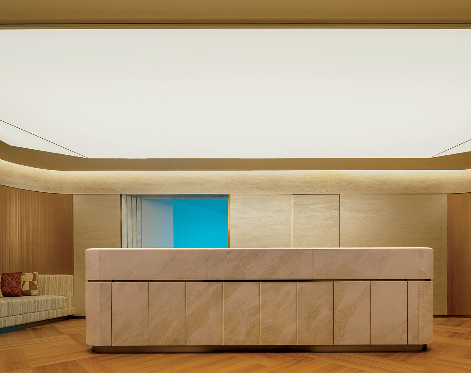 A modern, minimalist reception desk made of light-colored stone stands in a softly lit lobby with wood paneling and a cream sofa on the left.