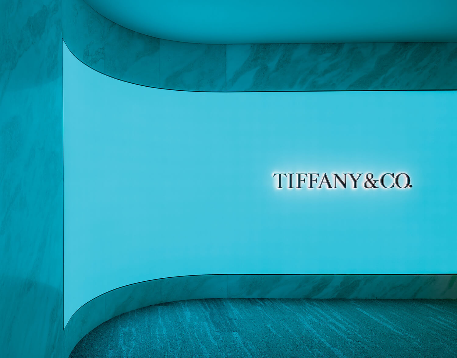 A curved, turquoise-lit wall with the illuminated Tiffany & Co. logo centered on it, set against a marble floor and background.