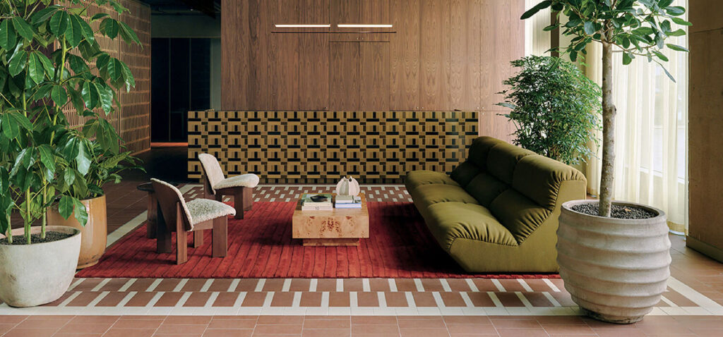 Modern lobby with a green sofa, two chairs, a wooden coffee table on a red rug, large potted plants, patterned front desk, and wood-paneled walls. Natural light enters from the right.